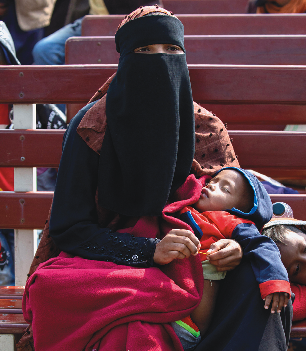 Fotografia 2. Mulher usando um lenço preto que cobre toda a cabeça e rosto, deixando à mostra apenas os olhos. Ela segura um bebê dormindo no colo.