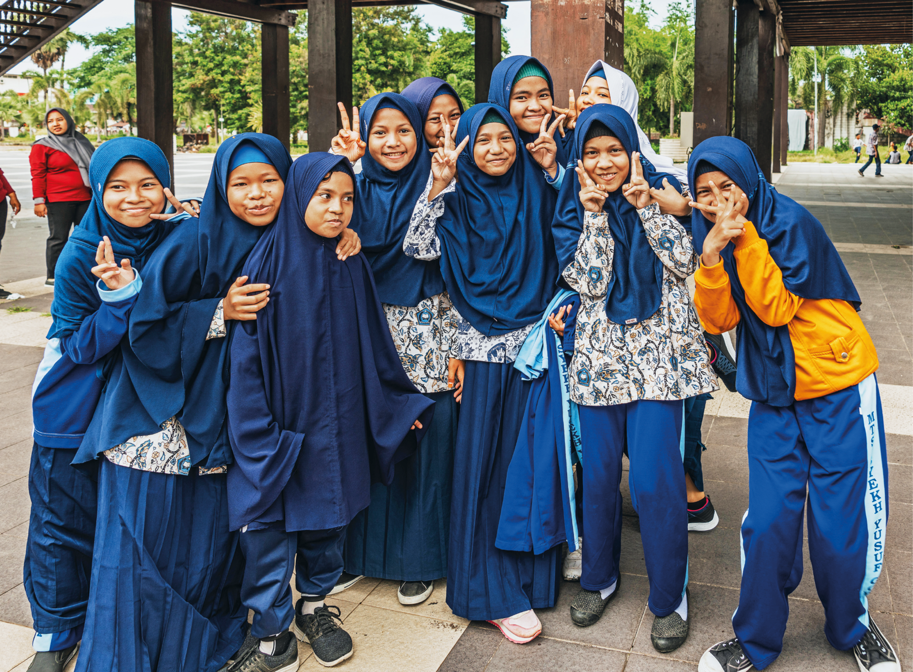 Fotografia. Meninas muçulmanas na escola. Usam lenços azuis para cobrir toda a cabeça e os ombros. Elas estão agrupadas, sorrindo para a foto.