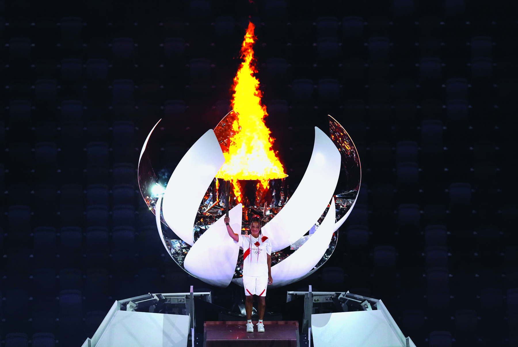 Fotografia. Mulher ergue tocha olímpica acesa. Ela veste bermuda e camiseta branca. Atrás dela a grande pira de fogo dentro de uma estrutura arredondada.