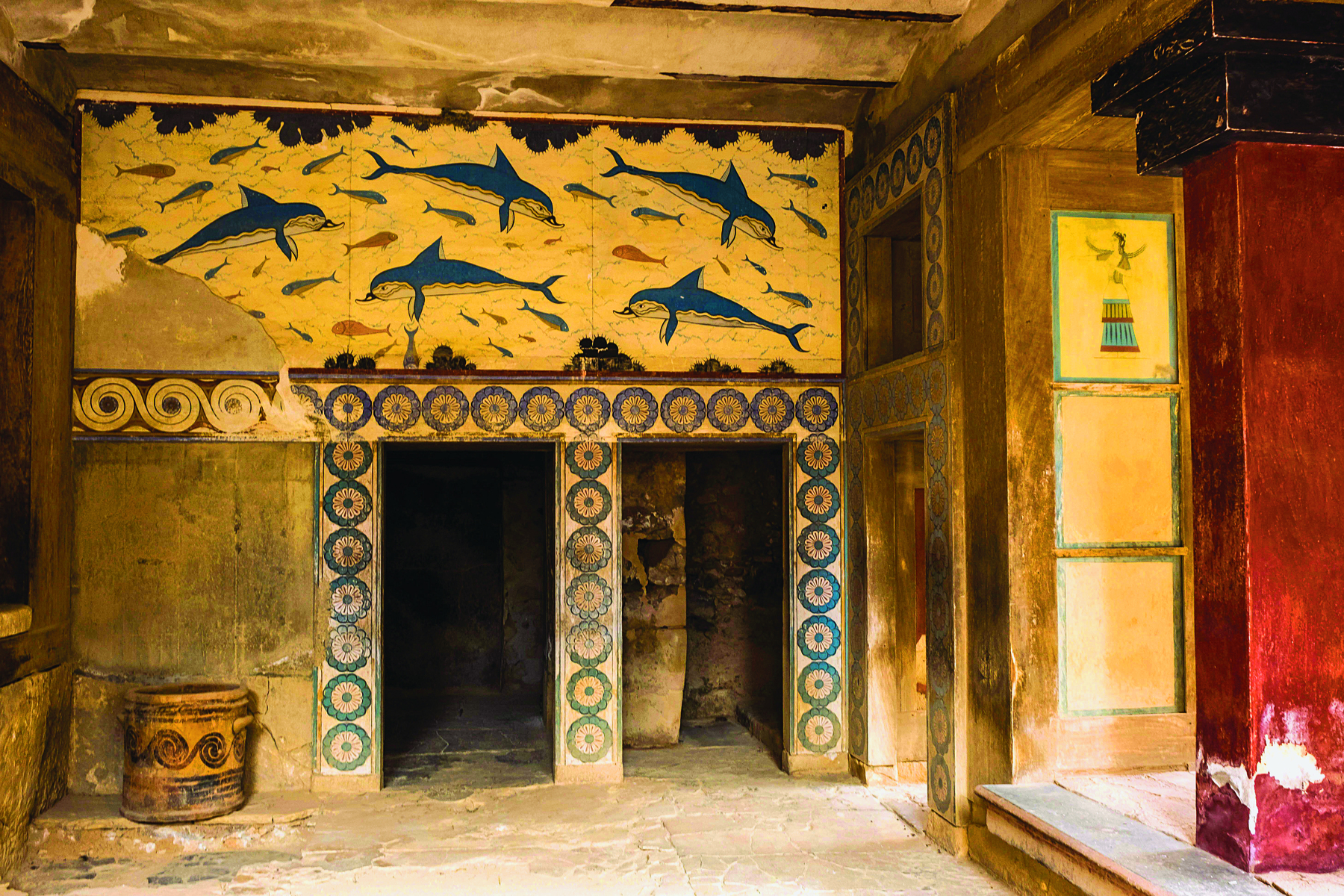 Fotografia. Destaque para a pintura de uma parede representando golfinhos azuis e outros peixes menores. A parte inferior esquerda da pintura está danificada. Abaixo da pintura principal, duas entradas decoradas com pinturas circulares azuis.