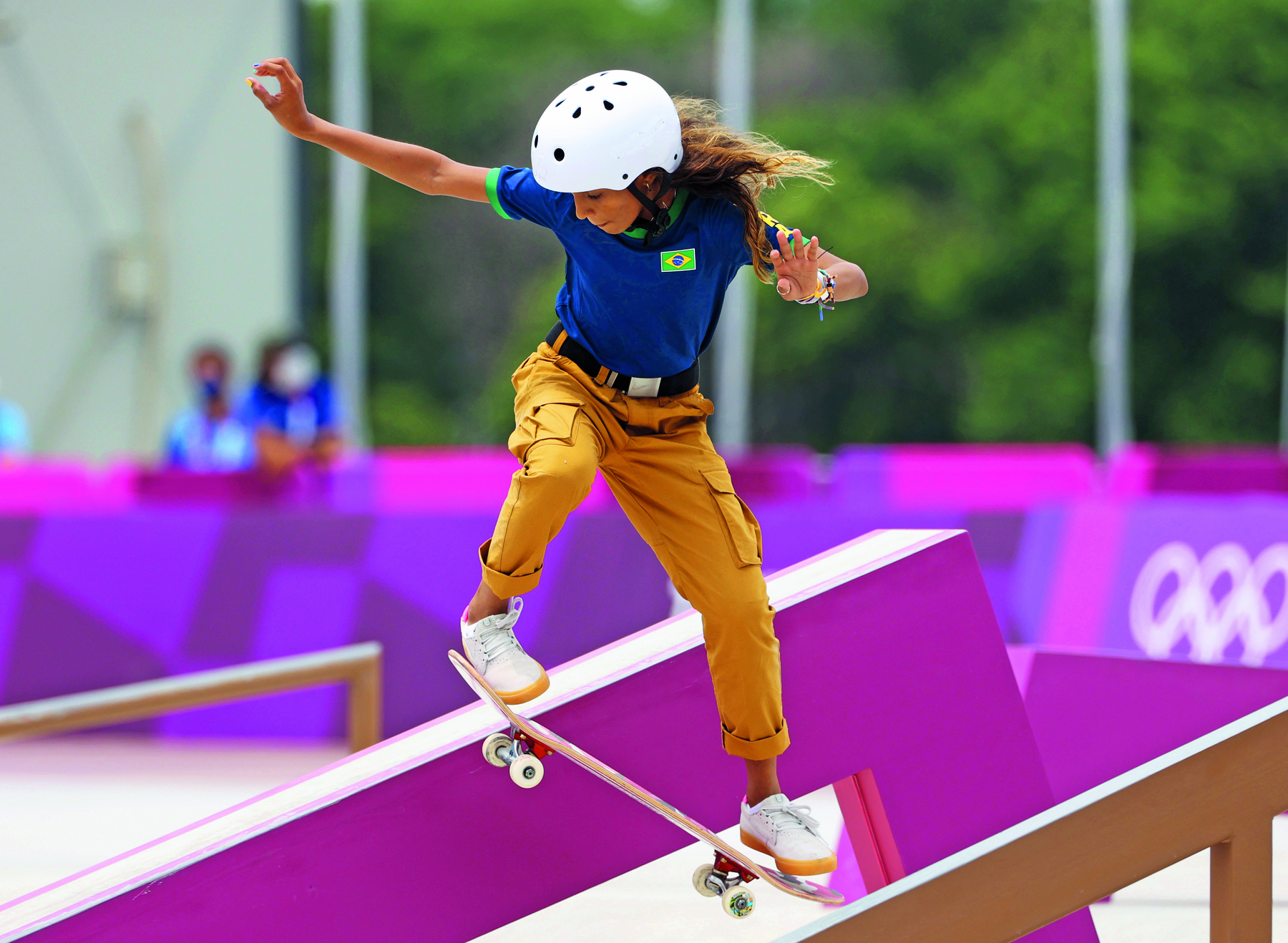 Fotografia. Uma menina realizando manobra sobre um skate. Ela veste camiseta azul com uma bandeira do Brasil desenhada, calça ocre dobrada na altura das canelas e um capacete branco na cabeça. Seus braços estão abertos, levemente flexionados. Os dois pés estão sobre o skate inclinado no ar.