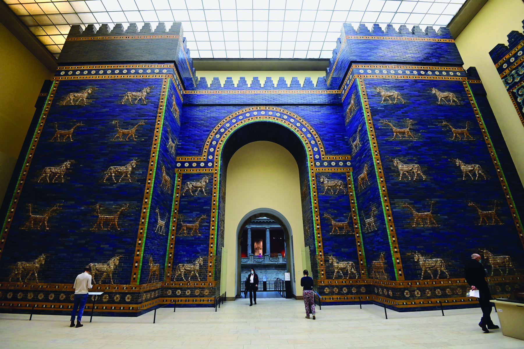 Fotografia. Pessoas observando um grande portal. Ele é formado por um grande muro azul, com animais desenhados em dourado. No centro, uma grande abertura em formato de arco.
