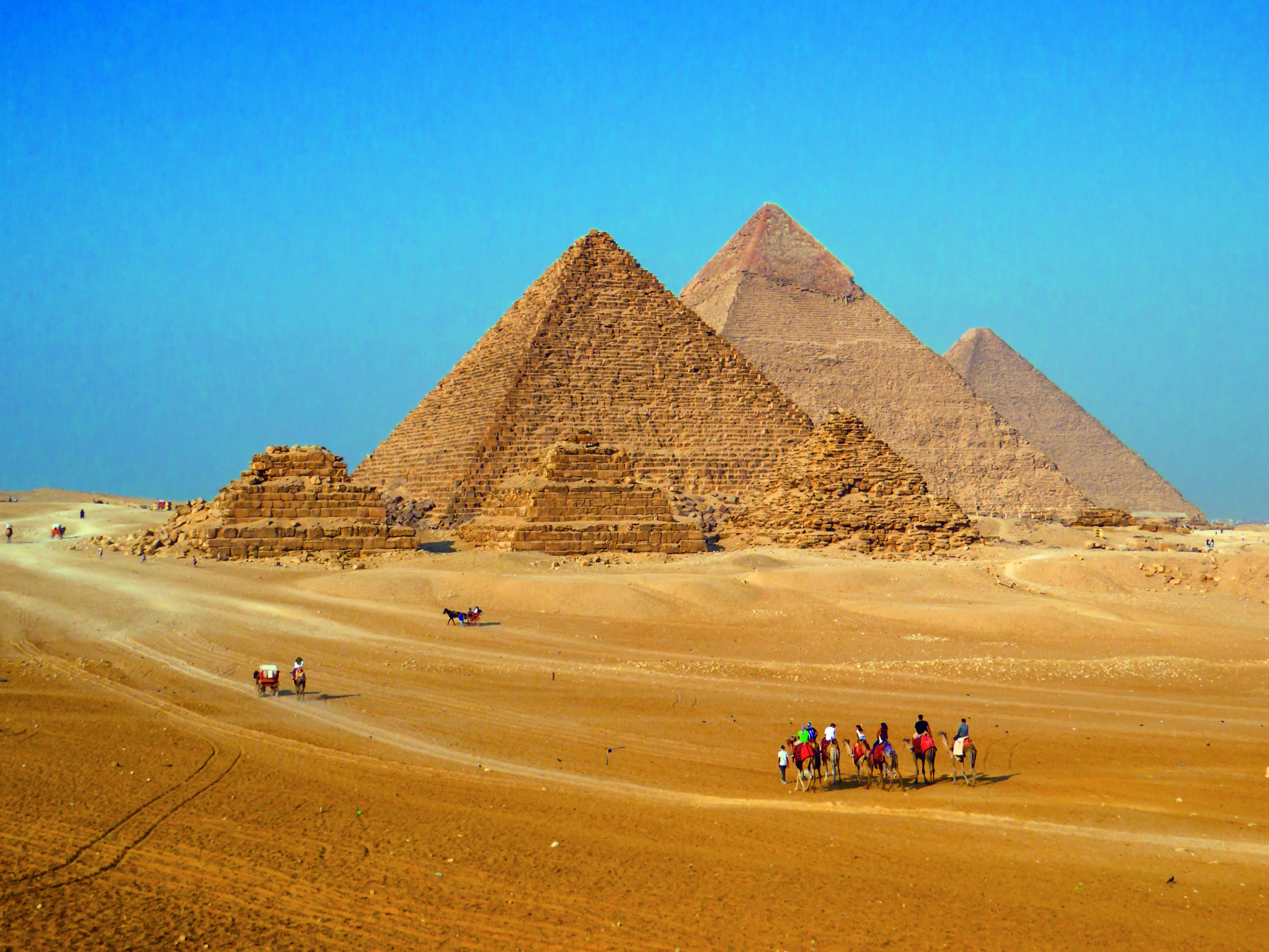 Fotografia. Paisagem desértica com seis pirâmides: três menores, na frente, e três maiores, ao fundo. As menores estão com suas estruturas danificadas. Na parte inferior da imagem, algumas pessoas montadas em camelos.