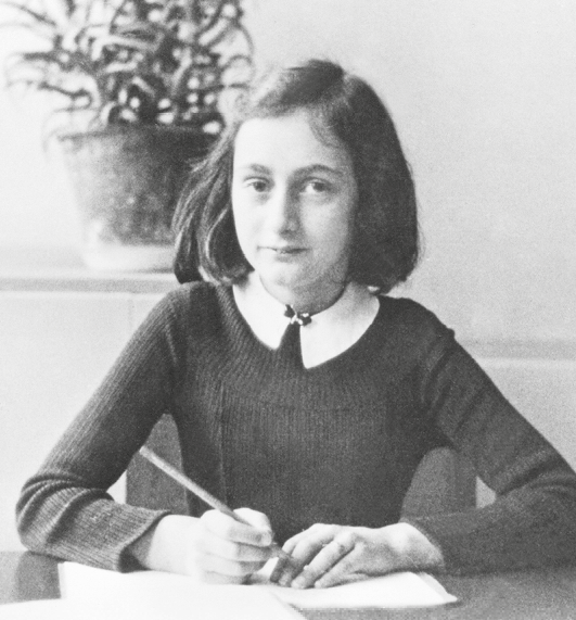 Fotografia em preto e branco. Anne Frank, garota de rosto afinado, pele branca, olhos escuros e arredondados. O cabelo é preto e liso na altura dos ombros. Ela usa uma camisa escura, de mangas compridas, e gola branca. Está sentada com os dois braços sobre a mesa, na qual há algumas folhas de papel. Em uma das mãos ela segura um lápis.