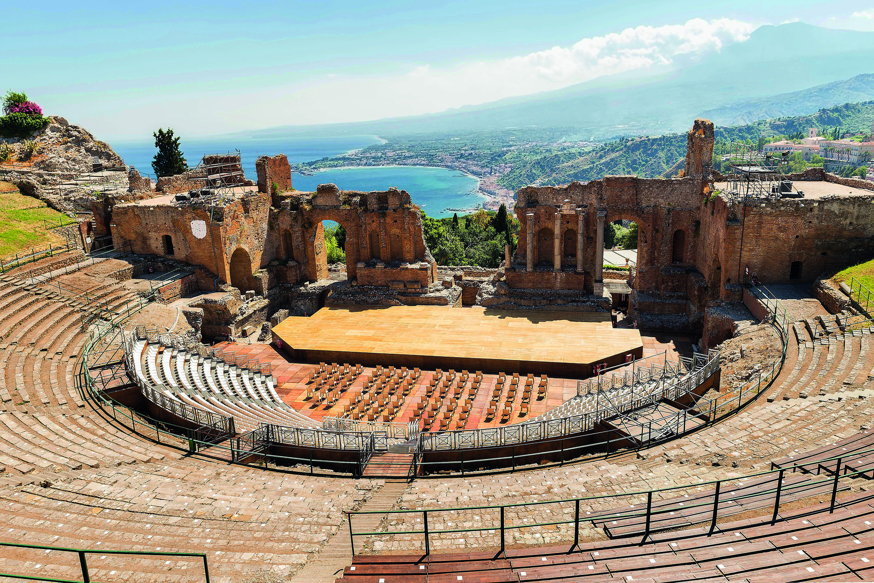 Fotografia. Teatro a céu aberto. Ele é oval e tem os assentos dispostos em grandes degraus. Na parte mais baixa, o palco. Atrás dele ruínas do muro composto por arcos.