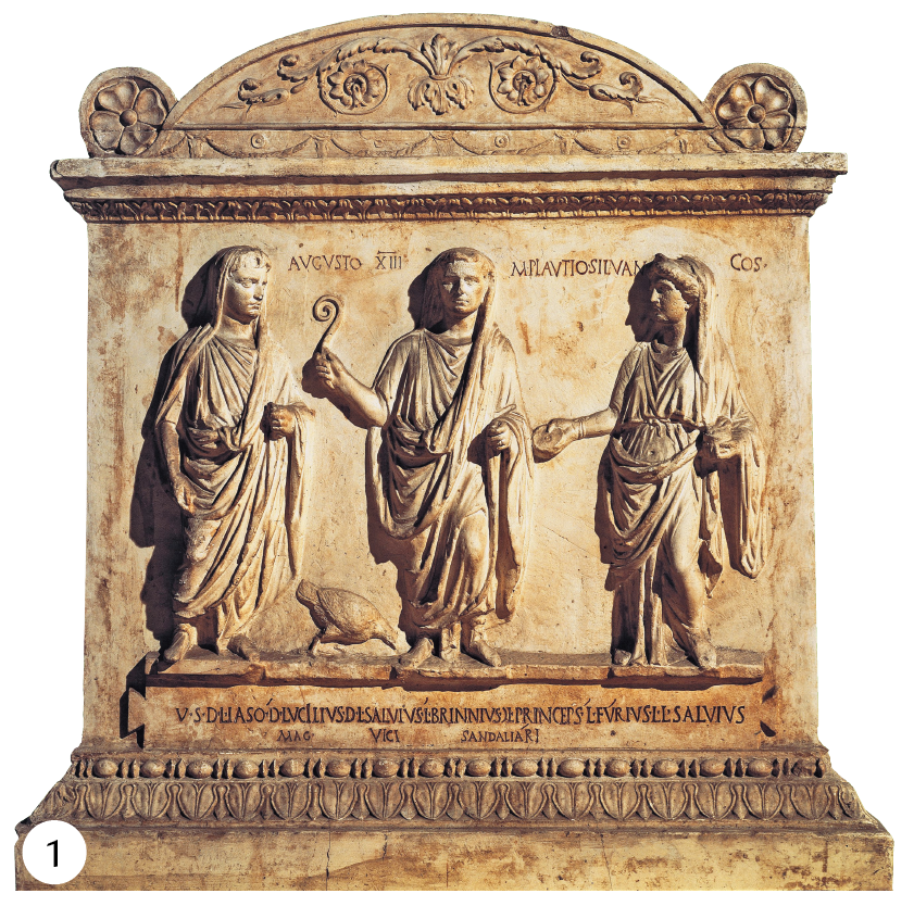 Fotografia 1. Escultura. Três pessoas sobre um altar. Todas vestem túnicas compridas. Na parte de baixo do altar inscrições. A escultura é toda decorada nas extremidades por arabescos.