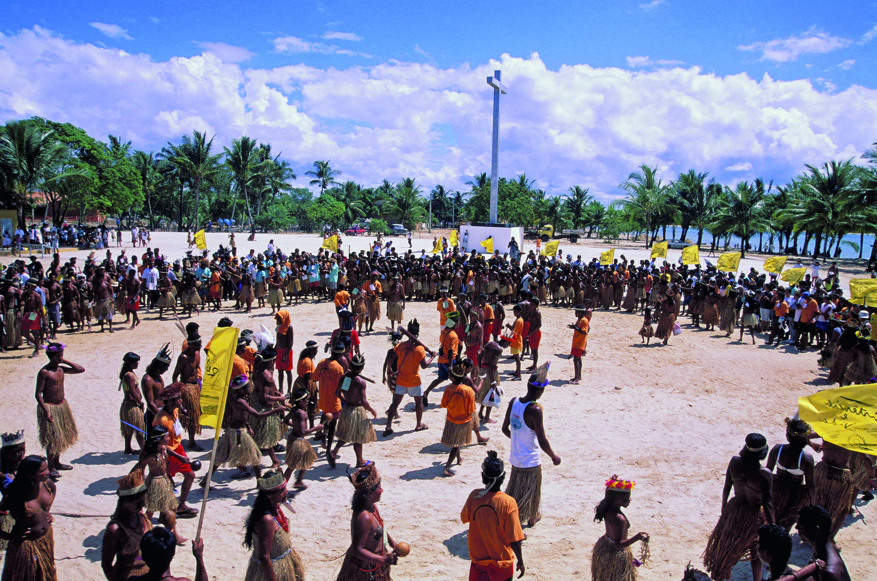 Fotografia. Grupo de pessoas indígenas reunidas em uma grande roda na praia. No centro, alguns indígenas de camiseta laranja.