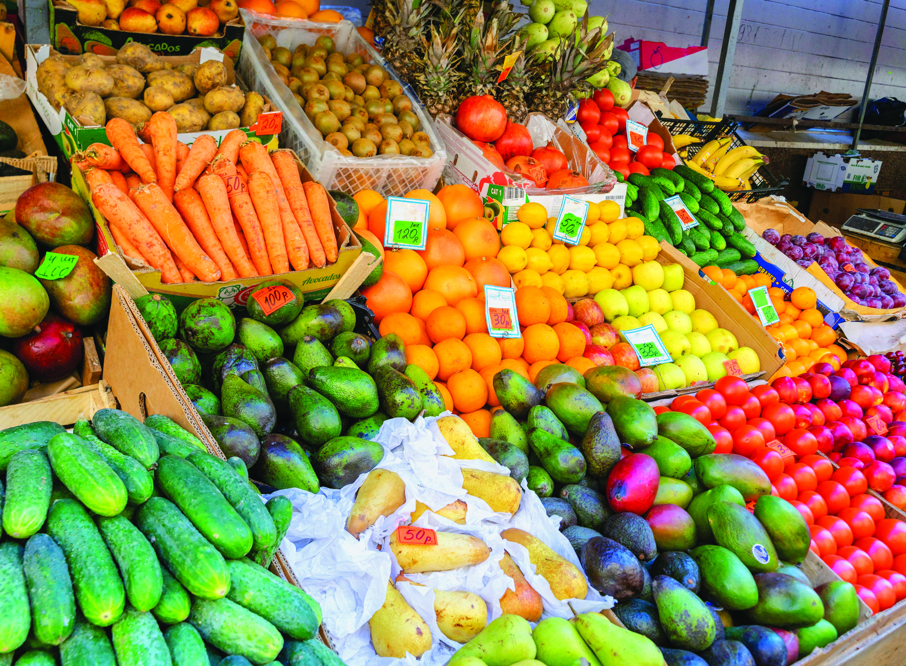 Fotografia. Frutas e legumes coloridos expostos em uma banca. Estão separados por tipo: mangas, pepinos, batatas, cenouras, abacates, peras, laranjas, abacaxis, tomates e bananas, entre outros.