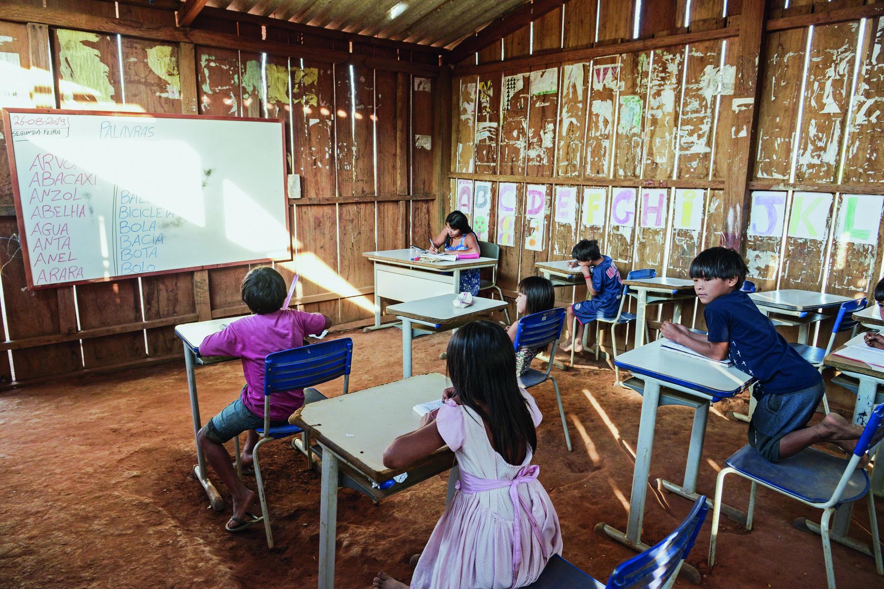 Fotografia. Sala de aula com o chão de terra e as paredes feitas com placas de madeira fina. Os alunos são crianças indígenas, e estão sentados em carteiras escolares. Estão de frente para uma lousa branca. No canto da sala, uma professora indígena sentada à mesa.