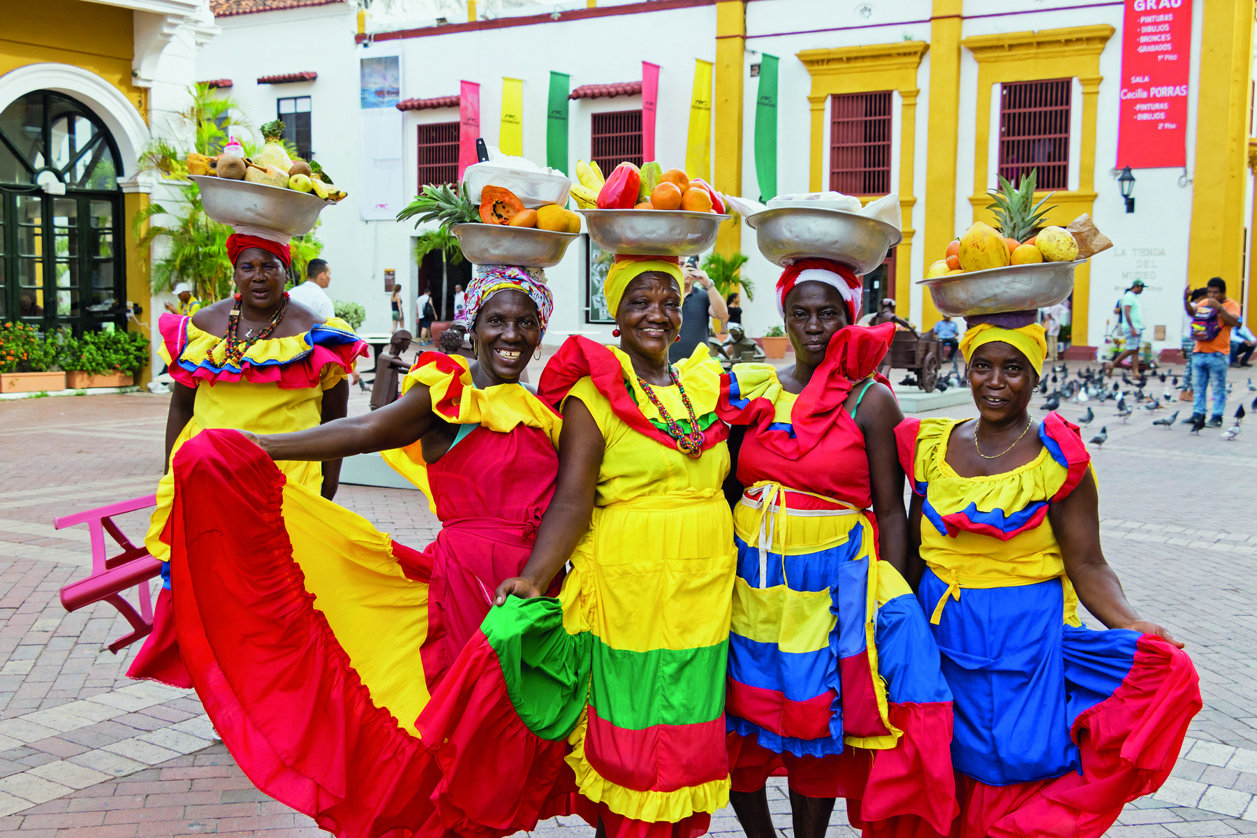Fotografia. Mulheres negras, sorridentes, posando para a foto. Usam vestidos coloridos em azul, vermelho, verde e amarelo. Todas carregam no topo da cabeça uma bacia de metal com frutas dentro.