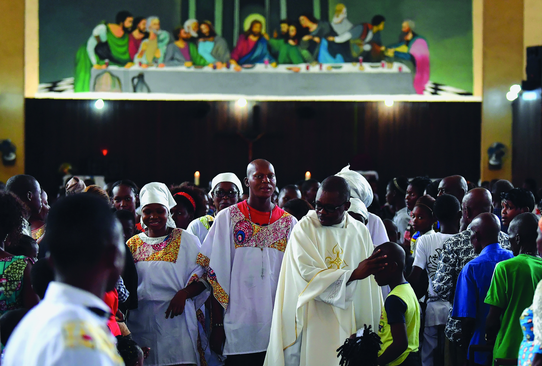 Fotografia. Pessoas reunidas dentro de uma igreja. A maioria é negra. O padre veste uma batina branca e usa óculos. Ao fundo, pintura de Jesus na mesa com os apóstolos.