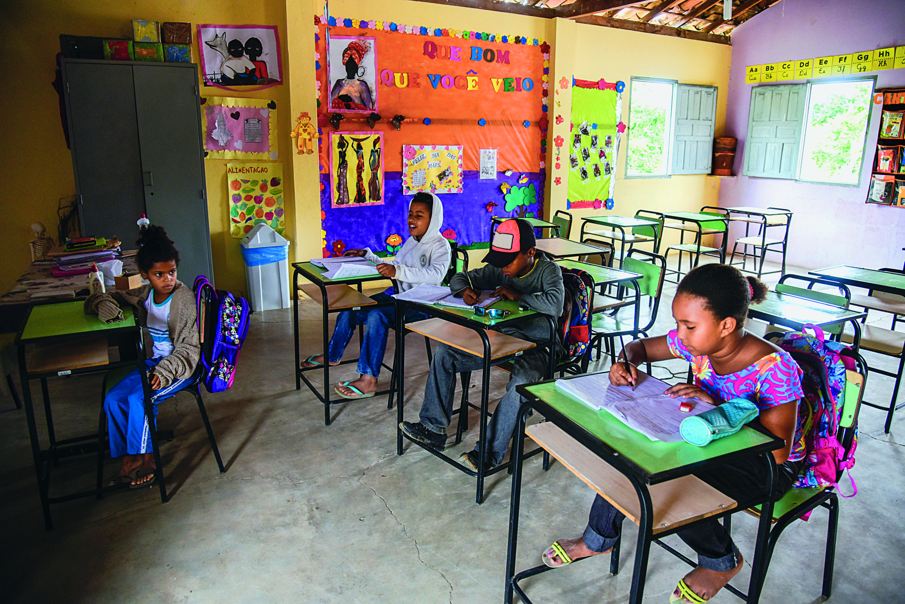 Fotografia. Meninos e meninas em uma sala de aula, sentados em carteiras escolares. As mochilas estão penduradas nas cadeiras. Ao fundo, parede decorada com vários cartazes coloridos.