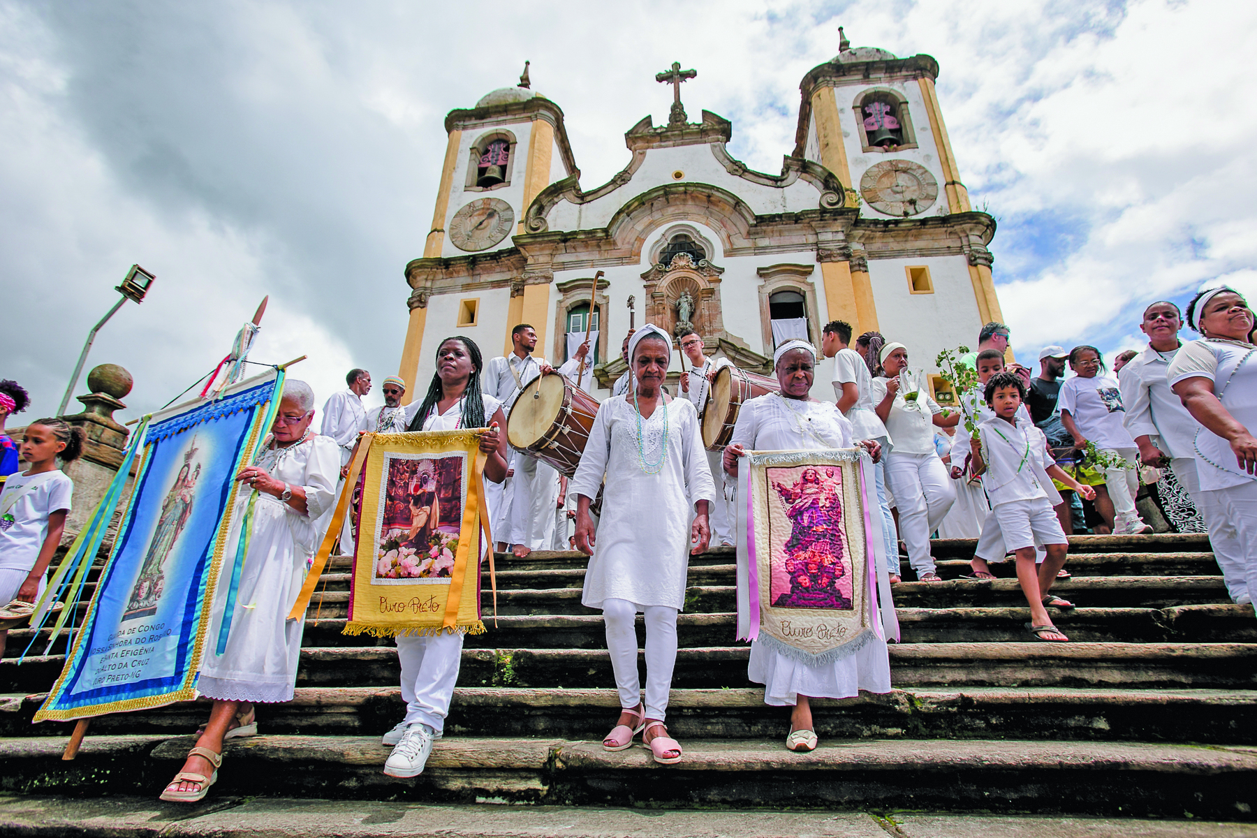 Fotografia. Pessoas vestidas de branco em uma escadaria em frente a uma igreja. À frente, quatro mulheres negras. Três delas carregam painéis com imagens de santos.