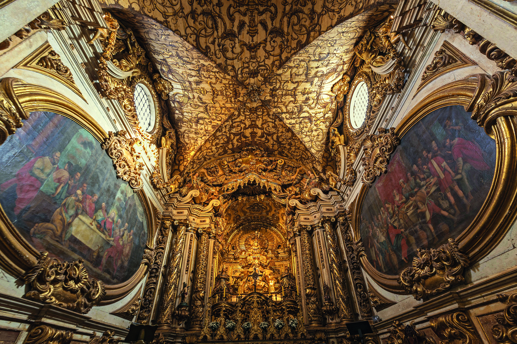 Fotografia. Vista parcial do interior de uma igreja. Nas paredes há grandes pinturas com molduras e ornamentos dourados. O teto é coberto por detalhes dourados. Ao fundo, altar ricamente decorado e grandes pilastras ornamentadas douradas.