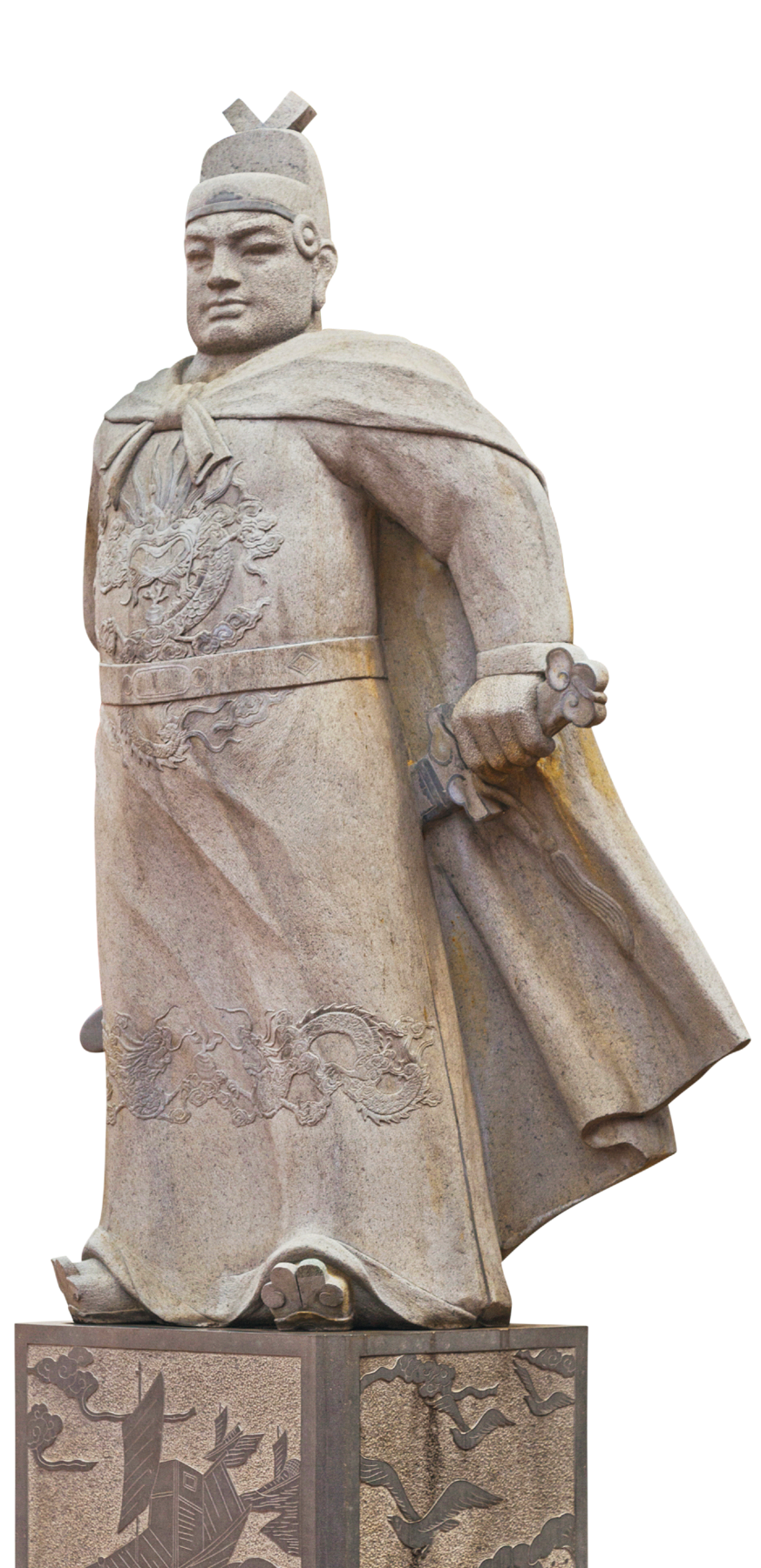 Escultura. Estátua em pedra de um homem com traços orientais. Tem o rosto largo e o queixo quadrado. Usa um chapéu e uma capa. Segura nas mãos uma espada, que fica entre a parte de trás de seu corpo e sua capa.