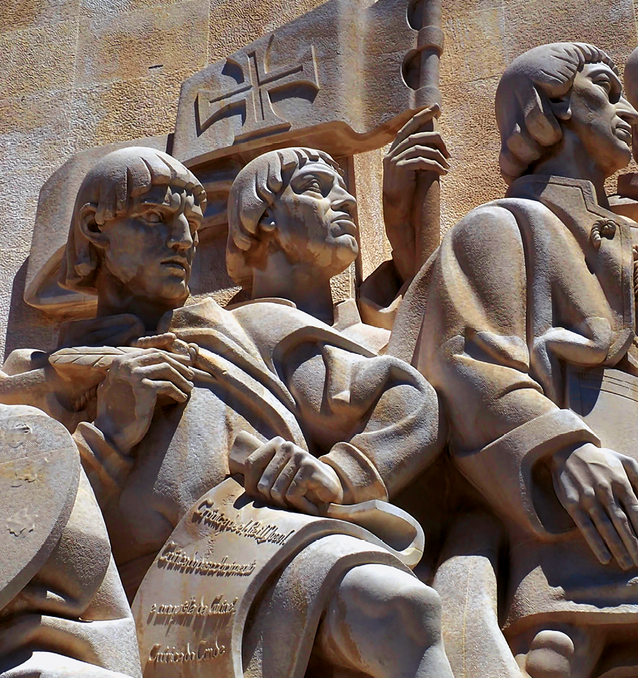 Fotografia. Detalhe de monumento de pedra composto por estátuas de homens aglomerados. Eles têm o cabelo liso na altura das orelhas. Atrás deles uma bandeira com uma cruz da Ordem de Cristo.