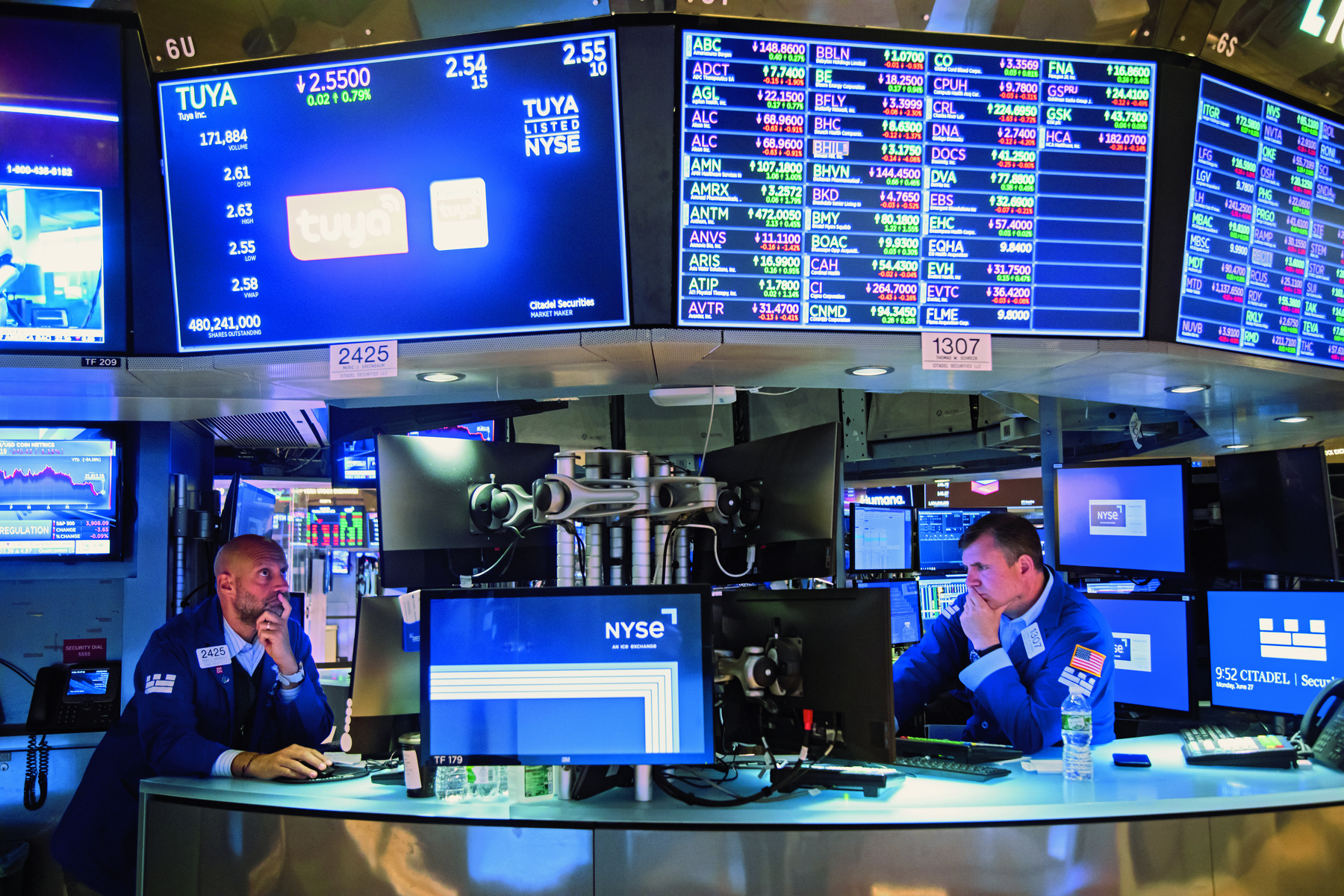 Fotografia. Dois homens sentados em cadeiras de encosto alto. Estão olhando para diversos monitores com informações financeiras. Os dois homens estão com uma mão na boca e a outra no mouse do computador. Ambos usam roupas azuis.