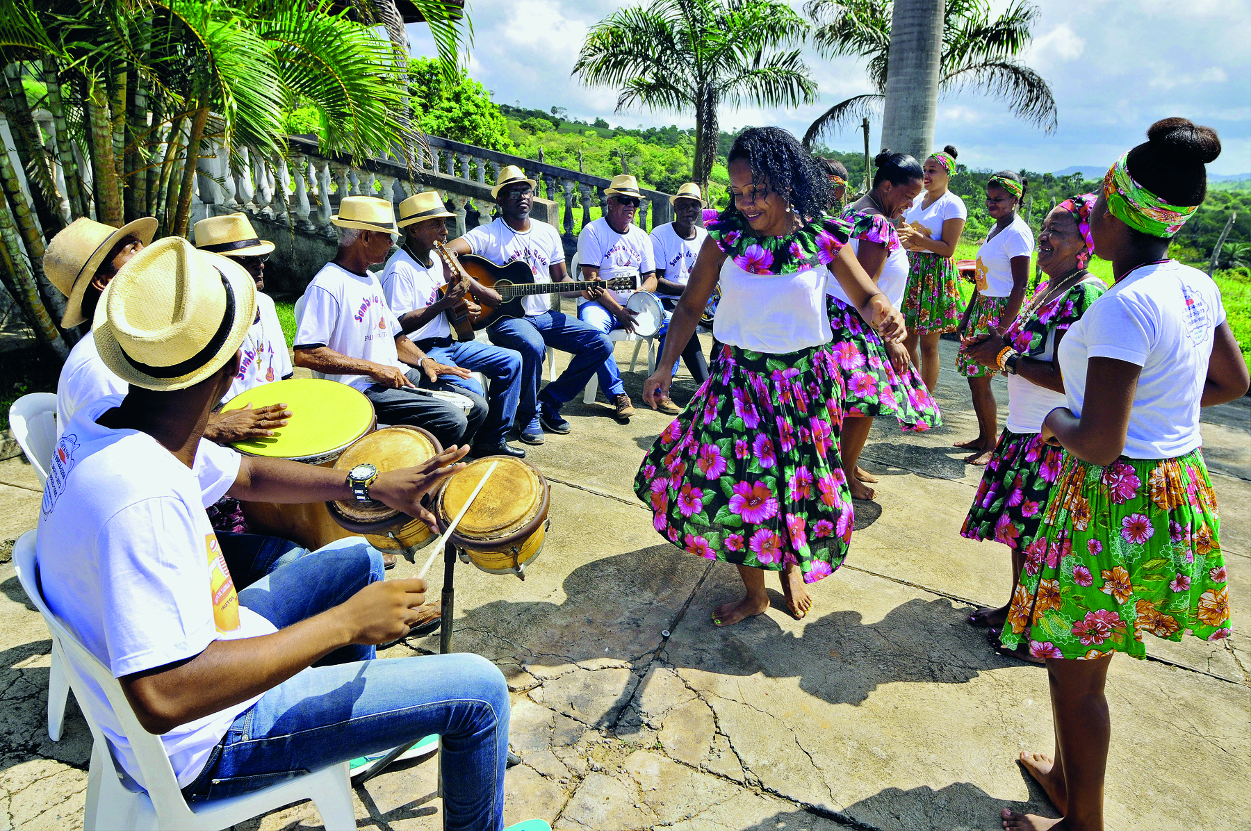 Fotografia. Grupo de mulheres negras de camiseta branca e saia florida, dançando. À esquerda, perto delas, homens negros de camiseta branca e chapéu de palha, sentados, tocando instrumentos musicais.