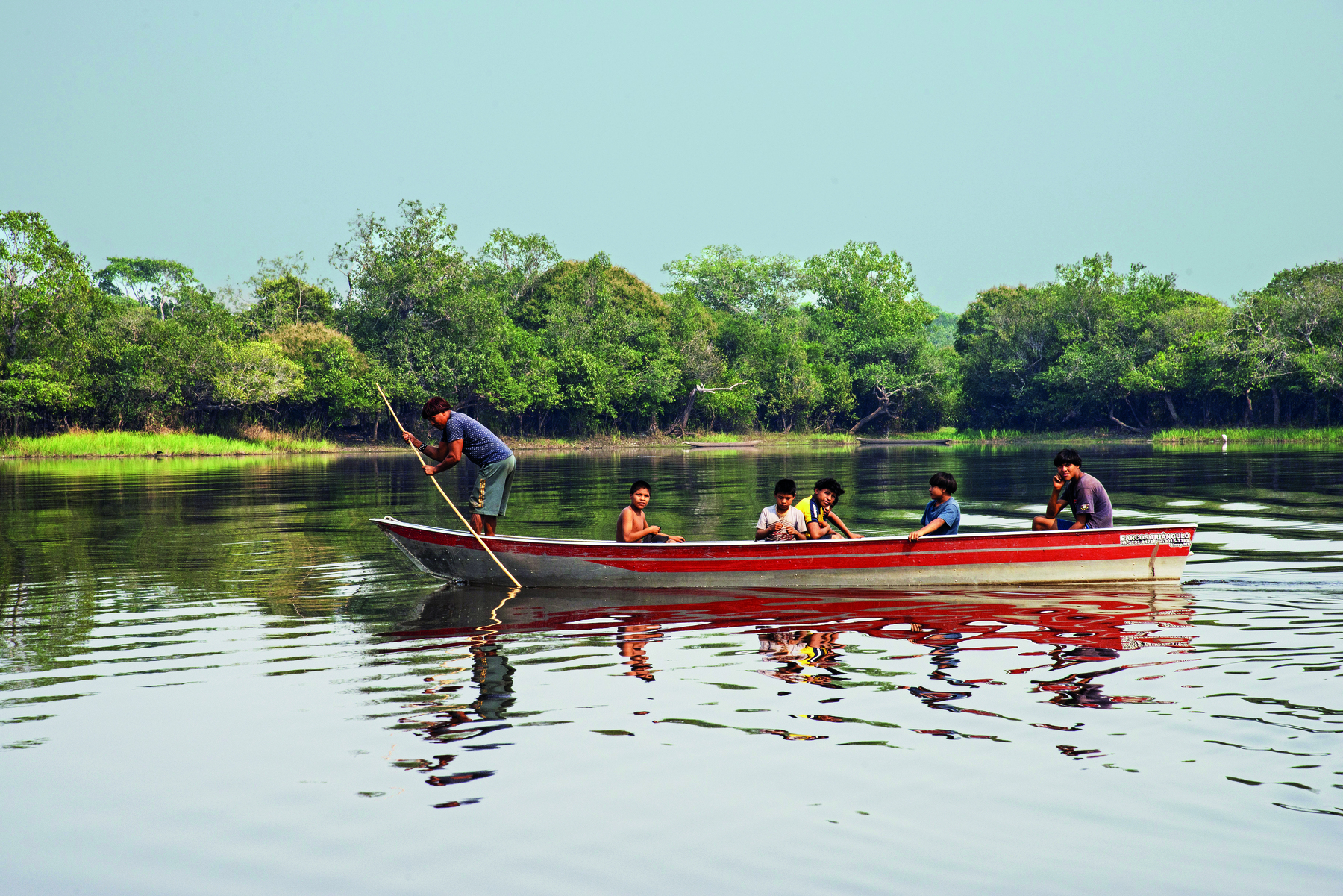 Fotografia. Pessoas indígenas navegando no rio em uma canoa. Um adulto está em pé, remando. Algumas crianças estão sentadas na embarcação.