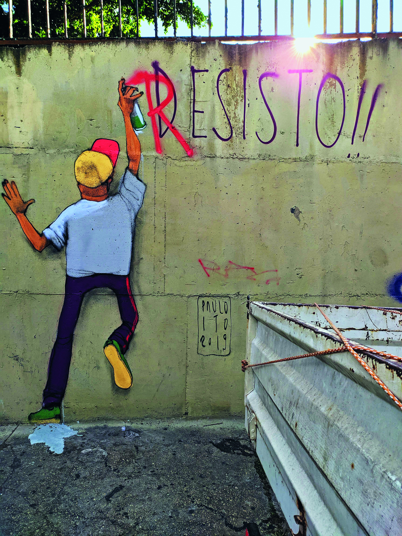 Fotografia. Grafite colorido em um muro de cimento representa uma pessoa de boné, de costas, escrevendo a letra R sobre o D da palavra DESISTO!!
