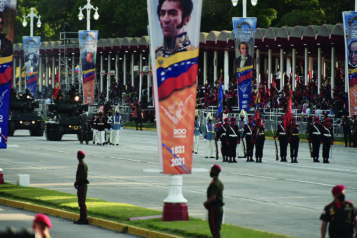 Fotografia. Grupo de militares desfila por uma avenida, alguns carregam bandeiras. Próximo a eles, tanques de guerra. Nas laterais, grandes banners com dados, fotos de alguns homens e bandeiras da Venezuela, com faixas amarela, azul e vermelha na horizontal, e um arco com estrelas brancas na faixa azul, no meio.
