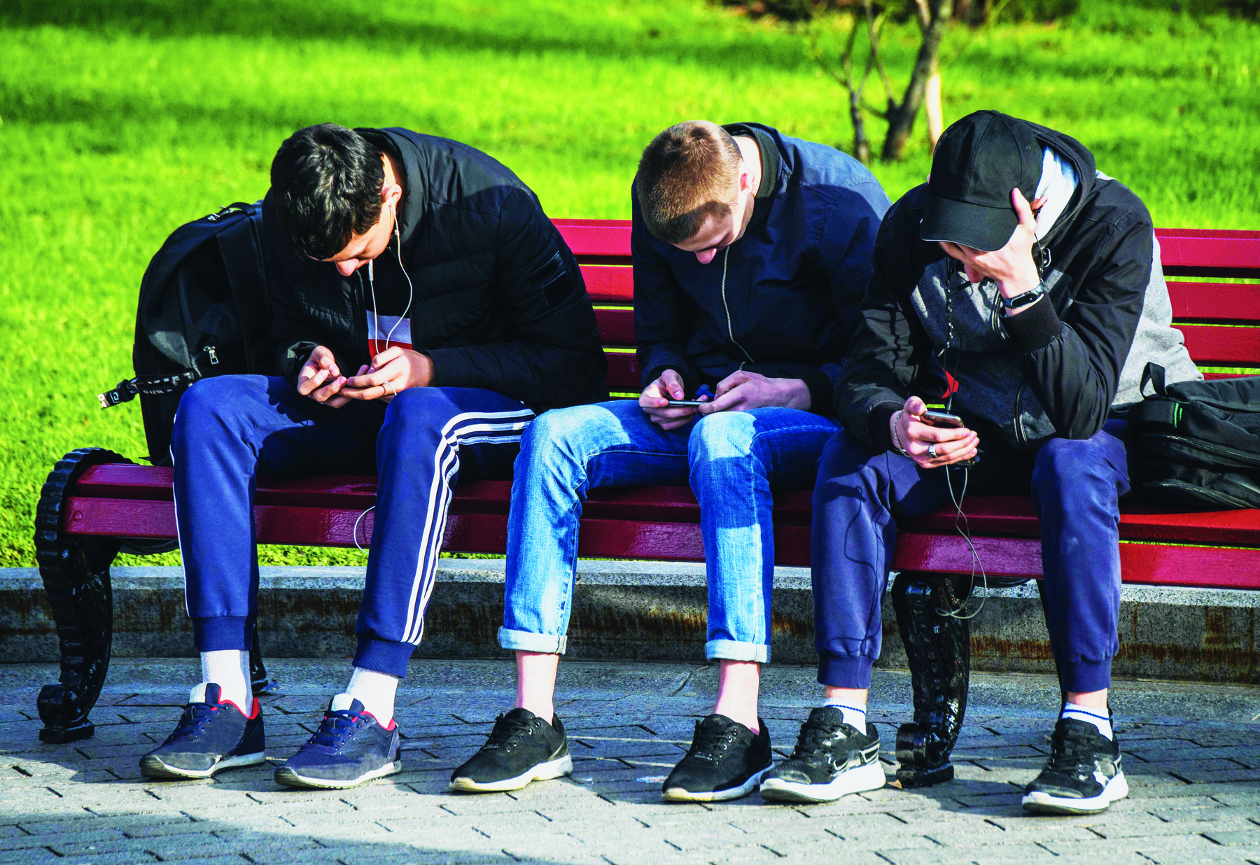 Fotografia. Três rapazes sentados em um banco vermelho. Estão um ao lado do outro, inclinados para frente, com a cabeça abaixada, cada um olhando um telefone celular. Ao fundo, um gramado.