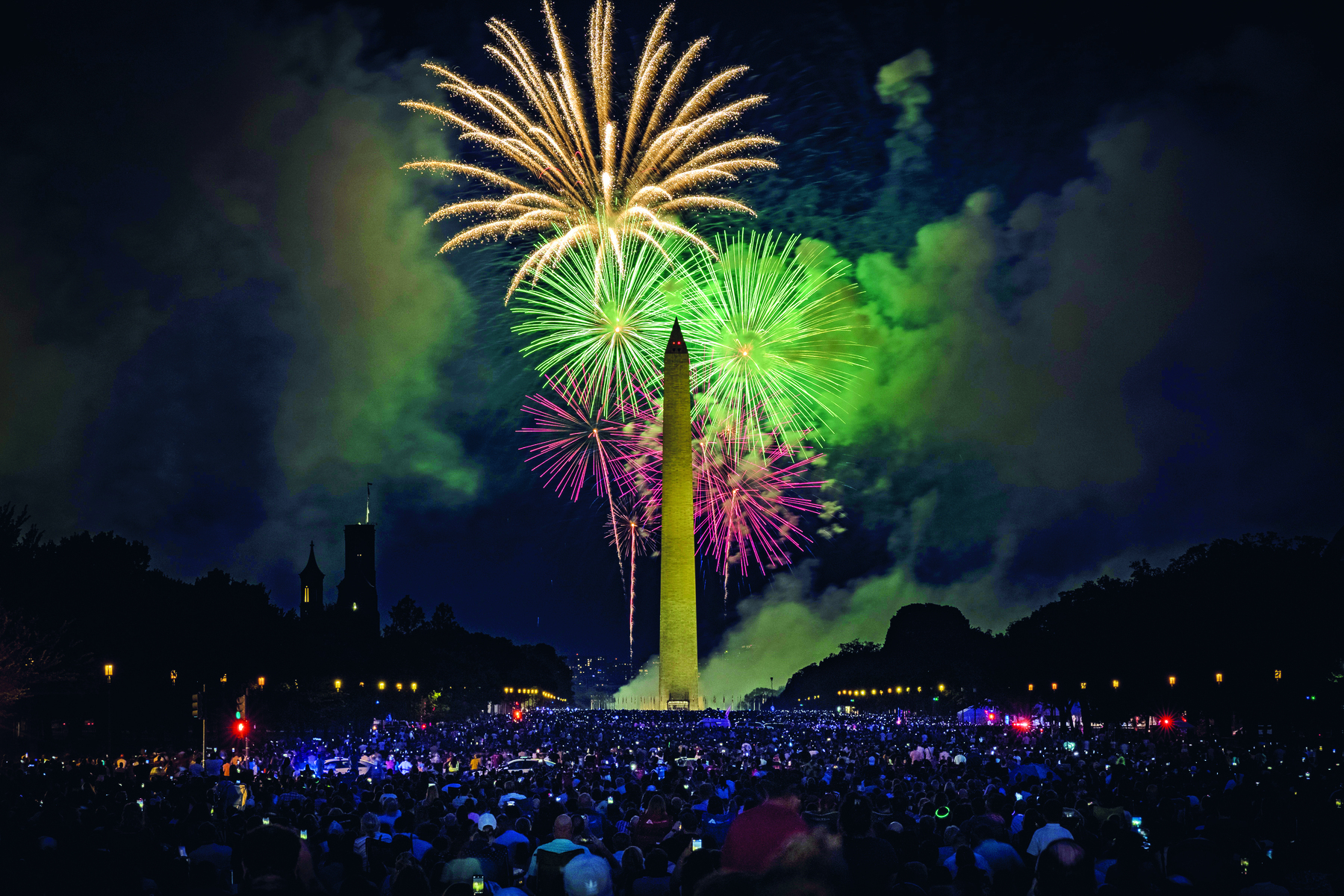 Fotografia. Multidão de pessoas assiste queima de fogos de artifício que colorem o céu noturno. Grandes feixes curvos e coloridos se espalhando pelo céu. No centro um obelisco, grande torre pontiaguda.