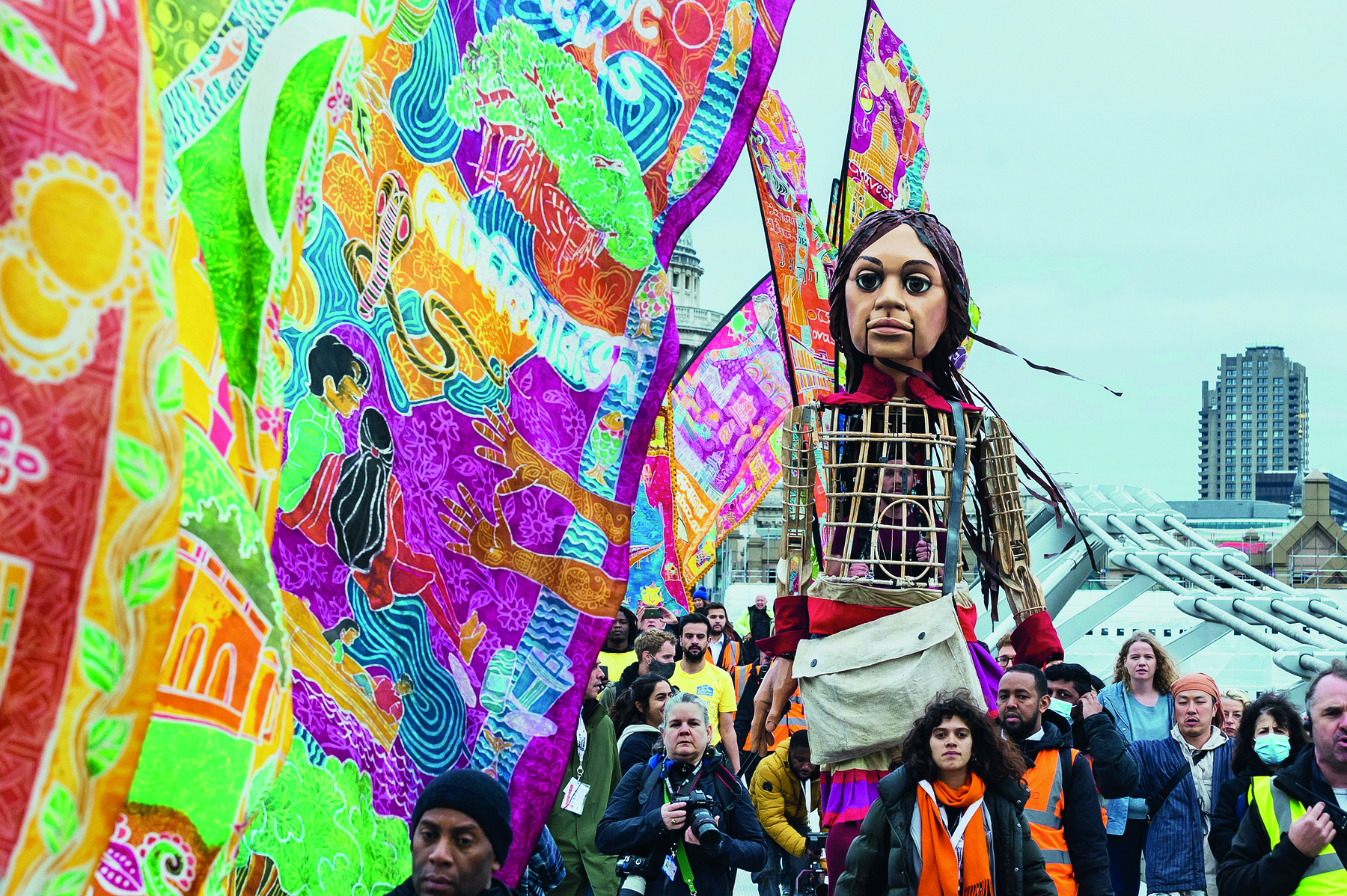 Imagem: Fotografia. Pessoas carregando um grande fantoche de uma criança. Ao redor, bandeiras com desenhos coloridos. Fim da imagem.