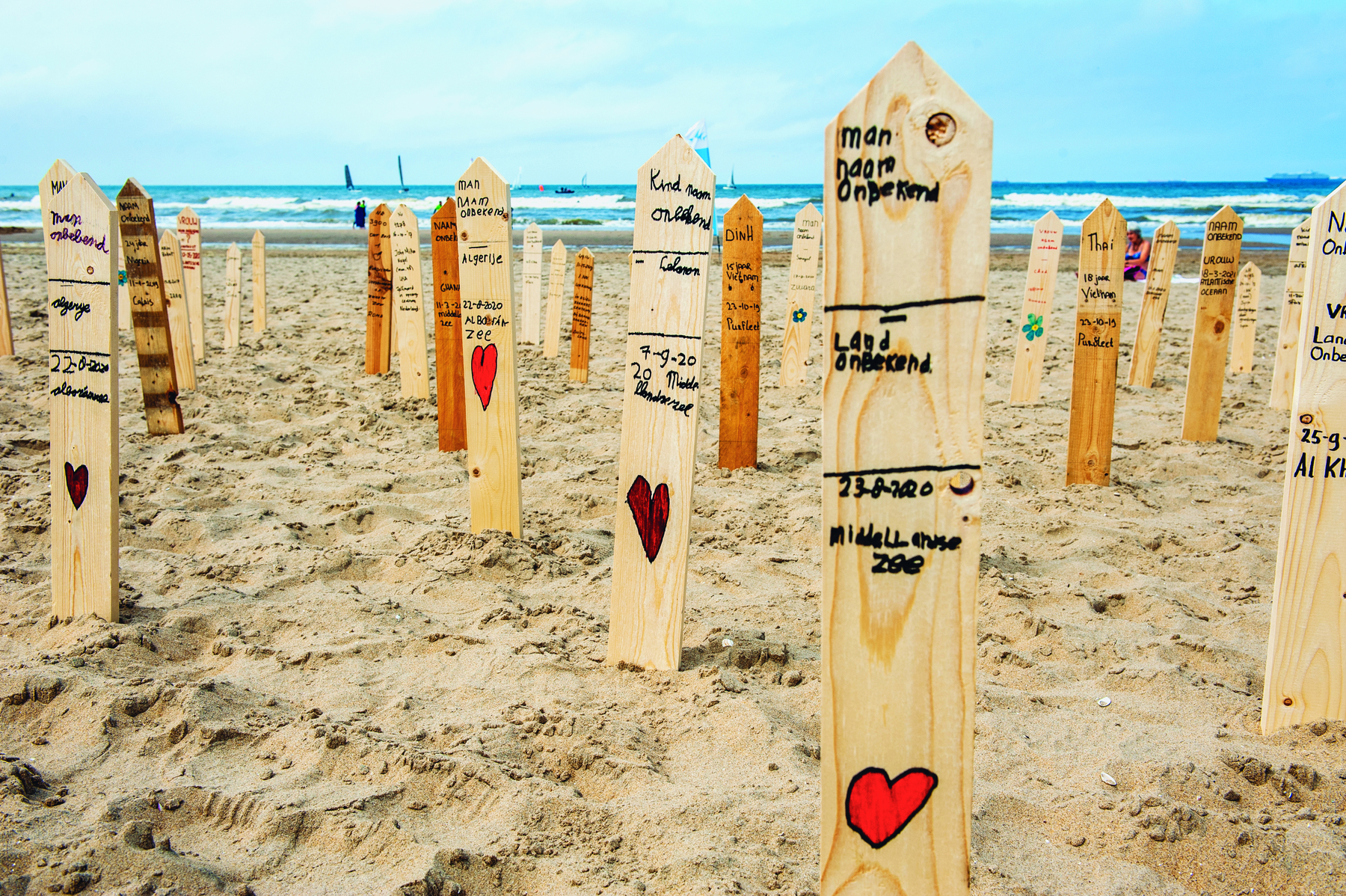 Imagem: Fotografia. Estacas de madeira fincadas na areia da praia. Em cada uma delas nomes escritos e um coração desenhado. Fim da imagem.