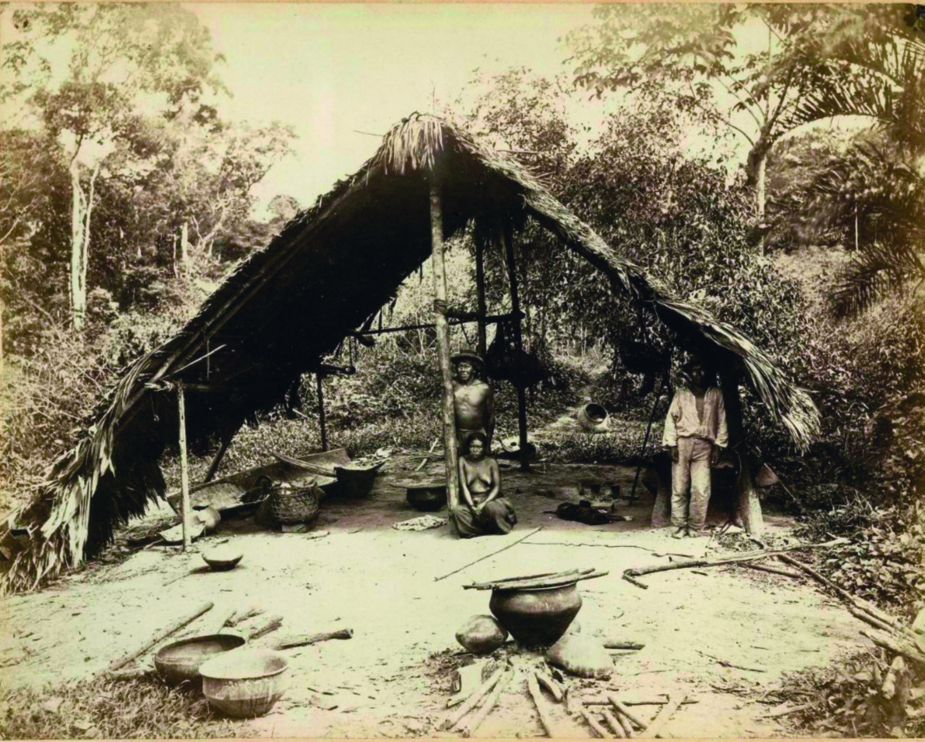 Fotografia em preto e branco. Indígenas dentro de uma cabana com teto triangular, feito de palha e madeira. Ao redor deles, pelo chão, muitos objetos, panelas e utensílios. Ao fundo, mata fechada.