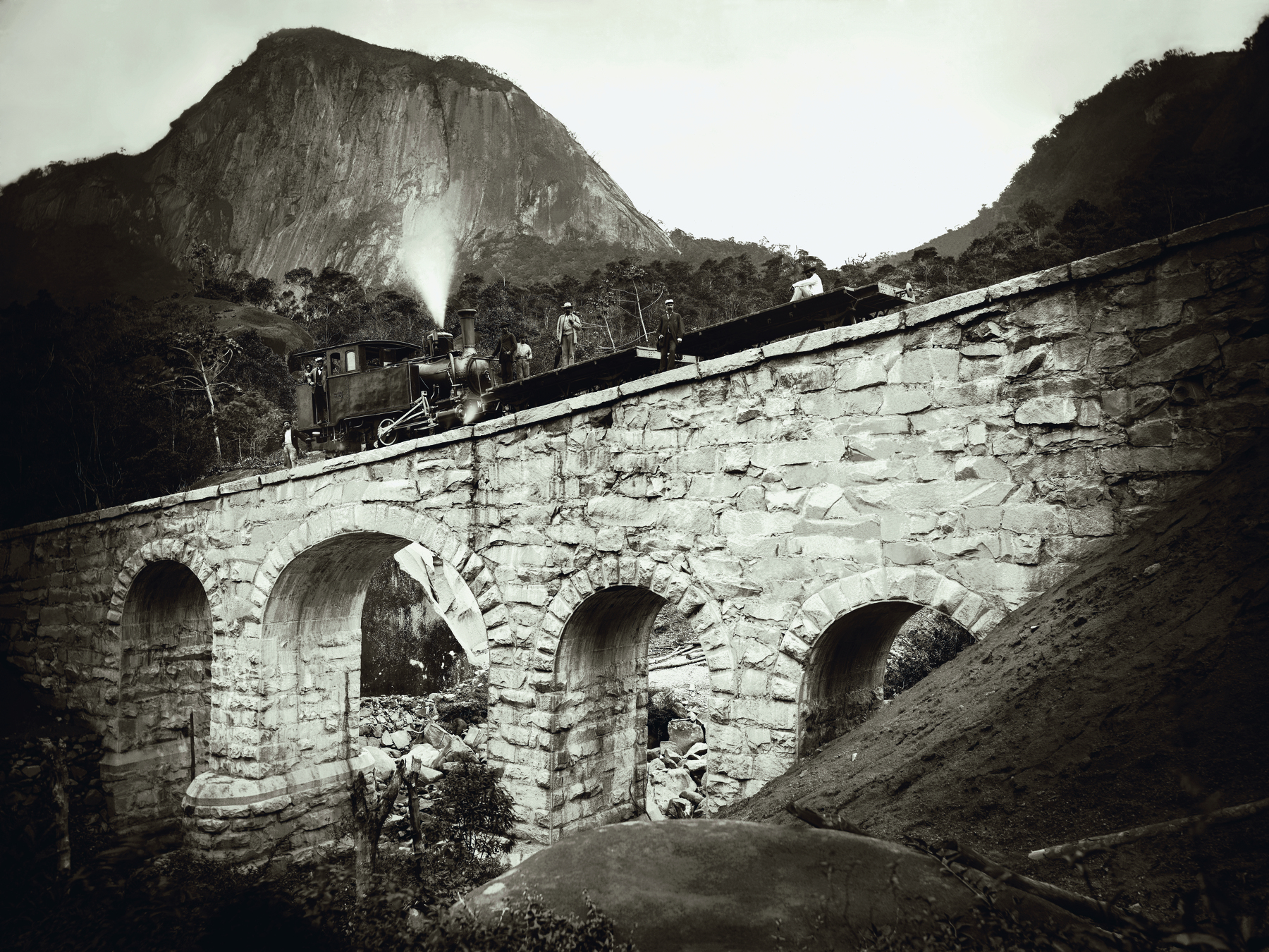 Fotografia em preto e branco. Grande ponte de pedras, com a base em arcos. No topo, uma estrada de ferro, um pequeno trem e pessoas sobre uma plataforma. Ao fundo, montanhas e a floresta.