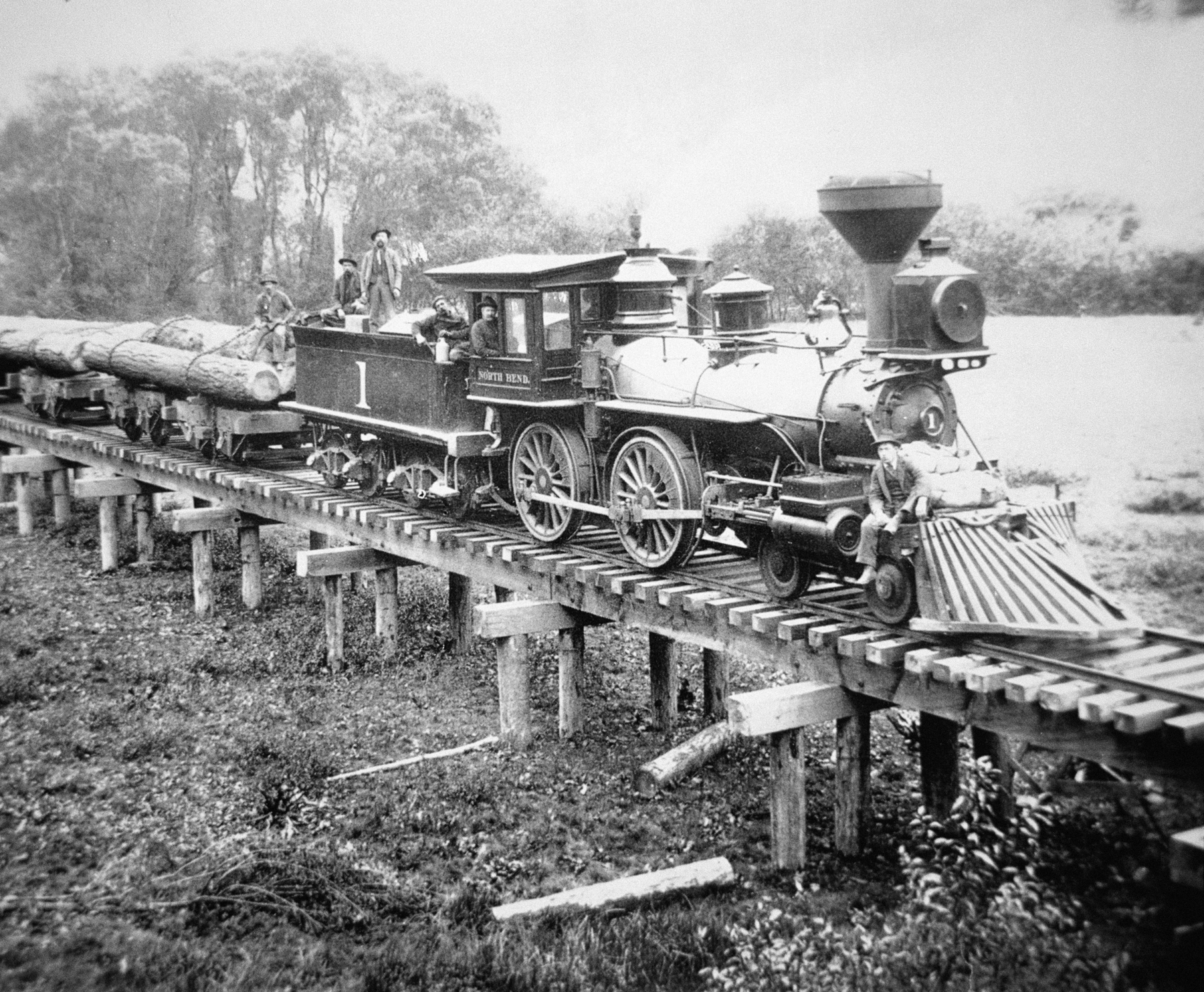 Fotografia em preto e branco. Trem carregando troncos de árvore passando por ferrovia suspensa em uma ponte de madeira. Na locomotiva, rodas grandes e uma chaminé.
