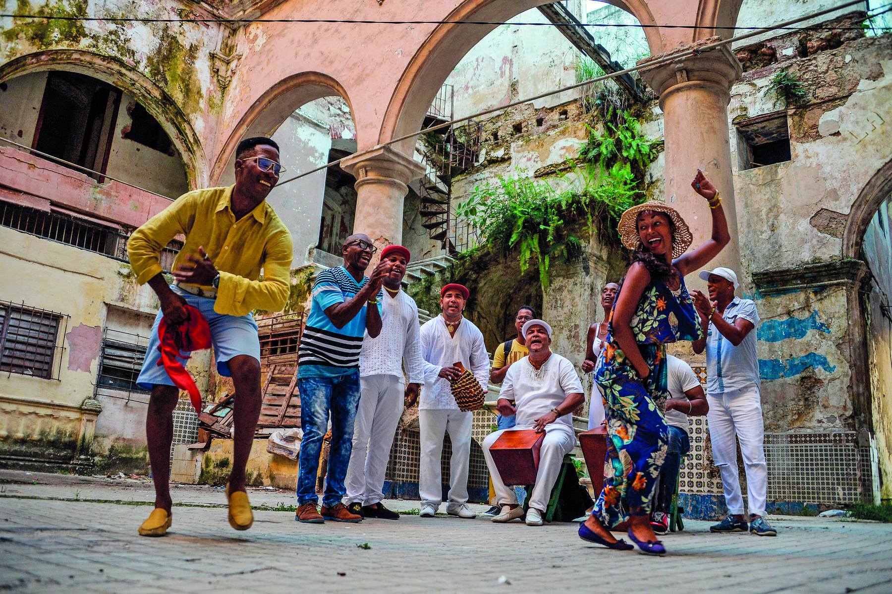 Fotografia. Grupo de pessoas tocando e dançando. Um homem de camisa amarela e bermuda jeans e uma mulher de vestido floral estão dançando. Atrás deles, homens cantando, alguns deles estão sentados tocando instrumentos.