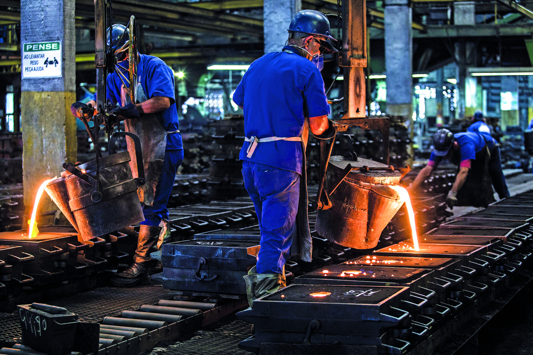 Fotografia. Homens trabalhando em uma metalúrgica. Eles vestem uniforme azul, botas, capacete, máscara, óculos de proteção e luvas. Eles despejam um líquido amarelado e luminoso dentro de moldes retangulares que estão no chão, dispostos em fila.