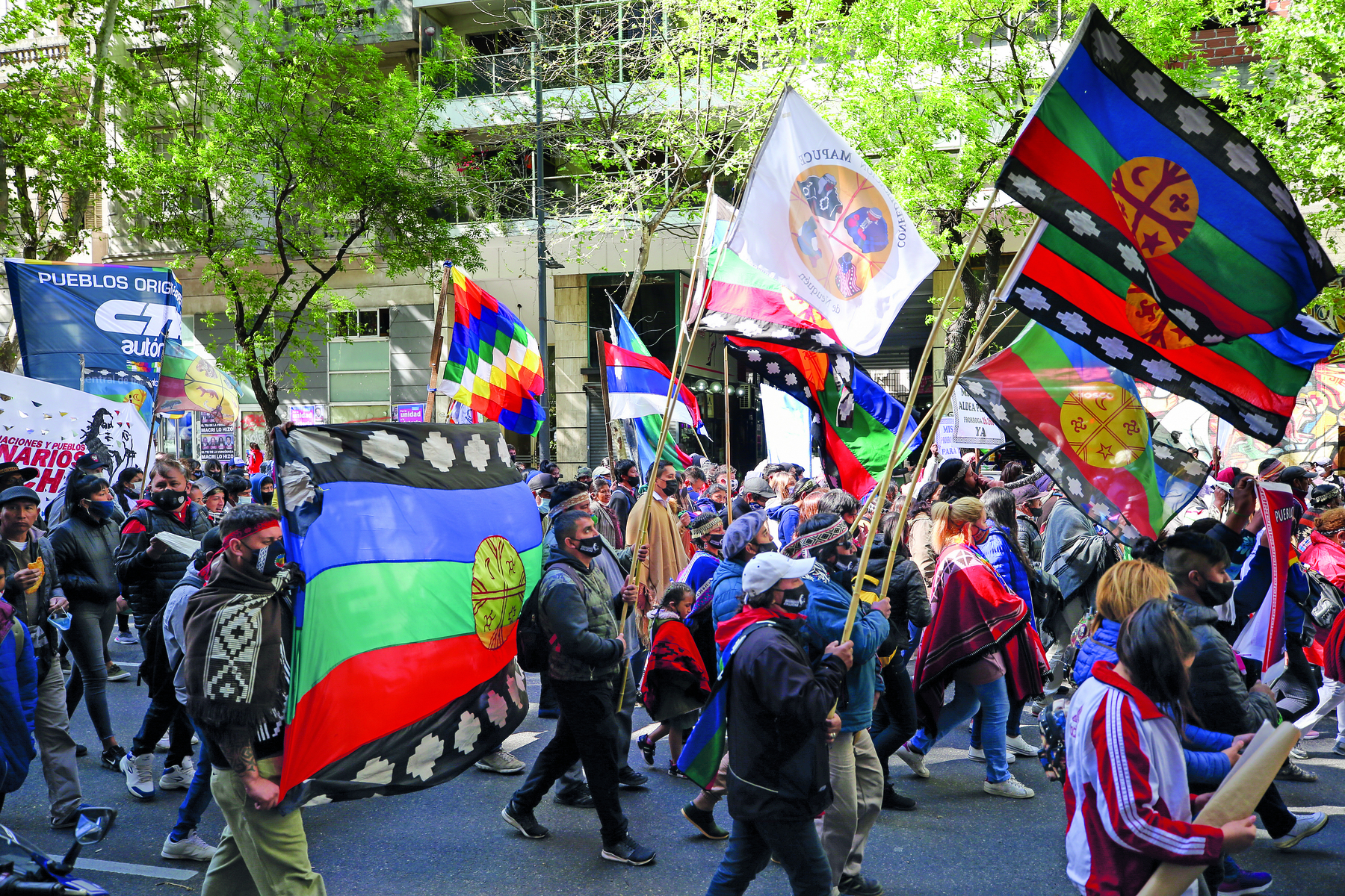 Fotografia. Muitas pessoas em uma manifestação na rua. Estão de máscara. Carregam bandeiras coloridas. Ao fundo, uma construção e algumas árvores.