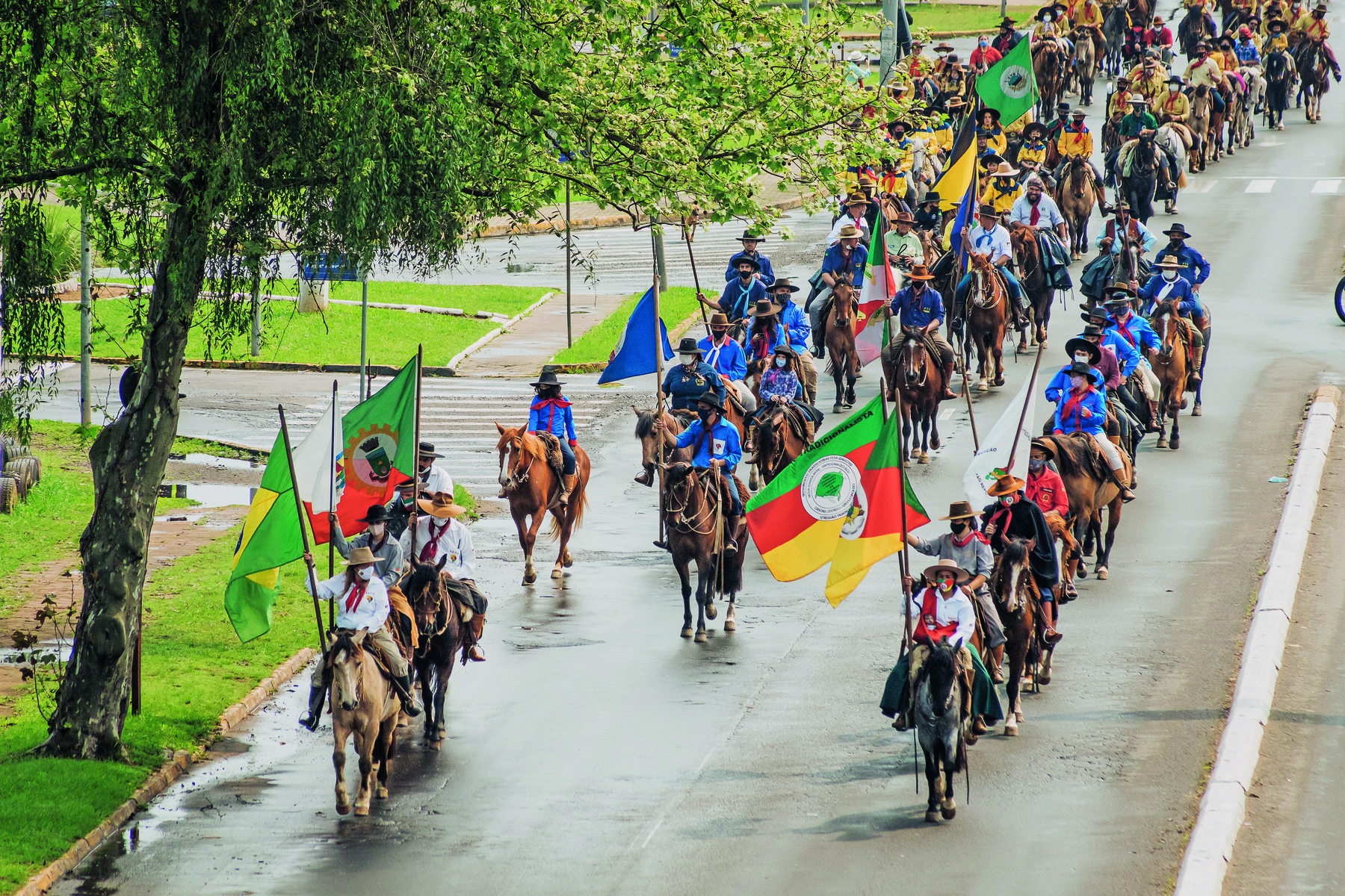 Fotografia. Pessoas andando a cavalo. Estão enfileiradas. Algumas usam chapéus e lenço vermelho no pescoço. As pessoas à frente trazem bandeiras do Brasil e do Rio Grande do Sul em mastros.