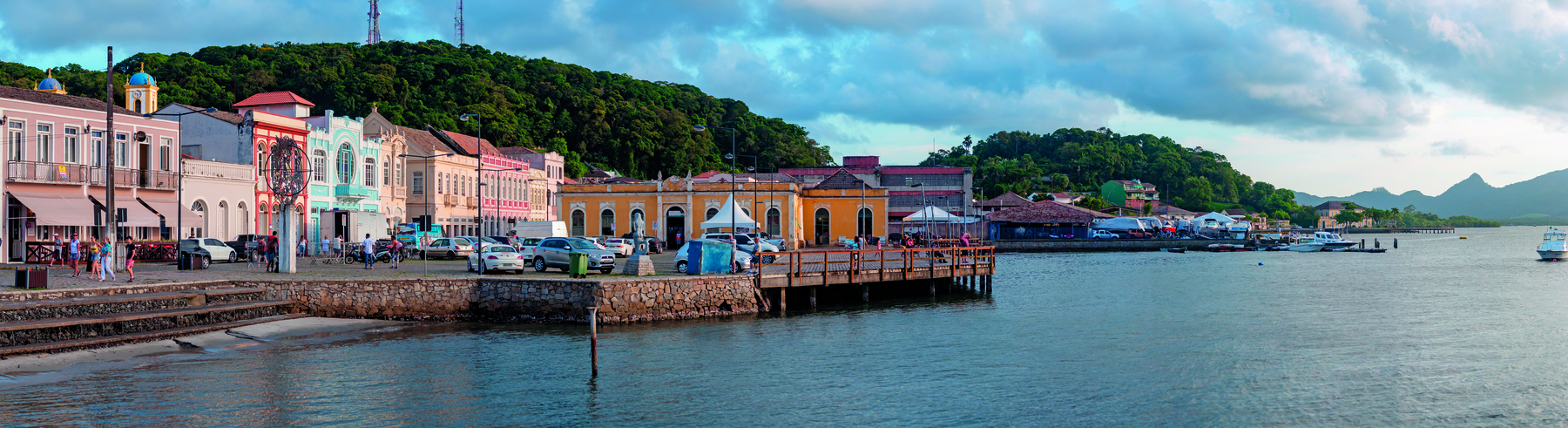 Imagem: Fotografia. À esquerda pequenos prédios coloridos e muita vegetação à beira mar. À direita, o mar. Fim da imagem.