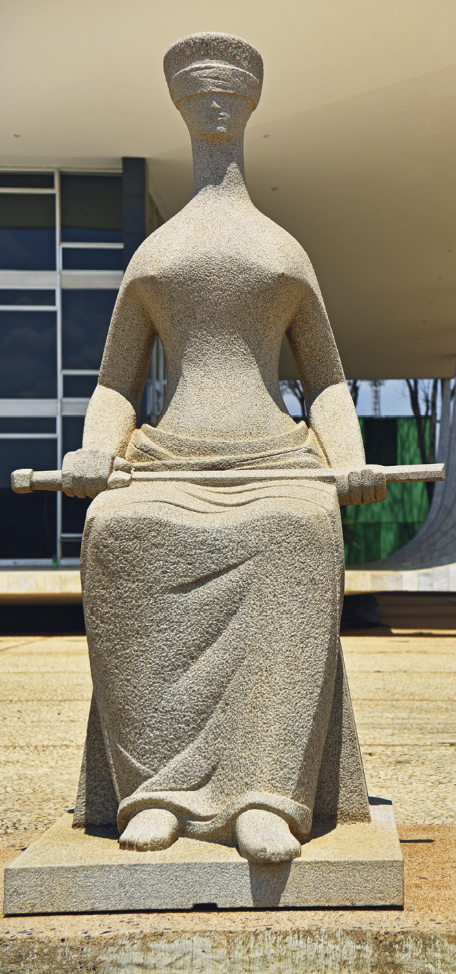 Fotografia. Escultura de uma mulher sentada com uma espada nas mãos, apoiada no colo. Um tecido grande e largo cobre a parte inferior do corpo, com exceção de seus pés. Tem o rosto fino e está de olhos vendados.
