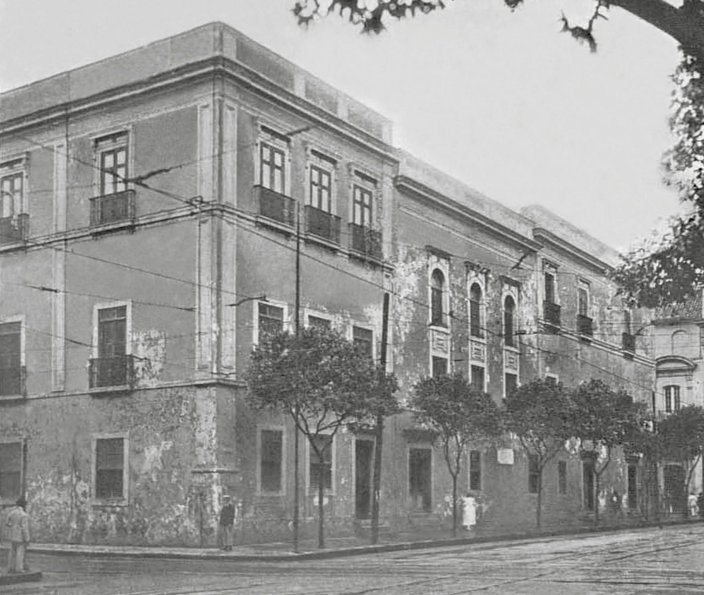 Fotografia em preto e branco. Edifício de esquina, com três andares e muitas janelas de moldura retangular. Na calçada algumas árvores.