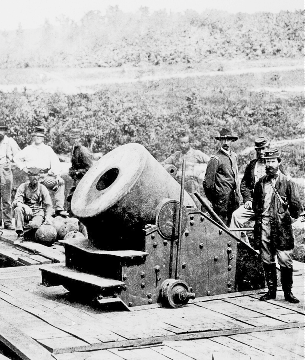 Fotografia em preto e branco. Homens de farda escura perto de um canhão grande e largo. Está preso em um tablado de madeira.