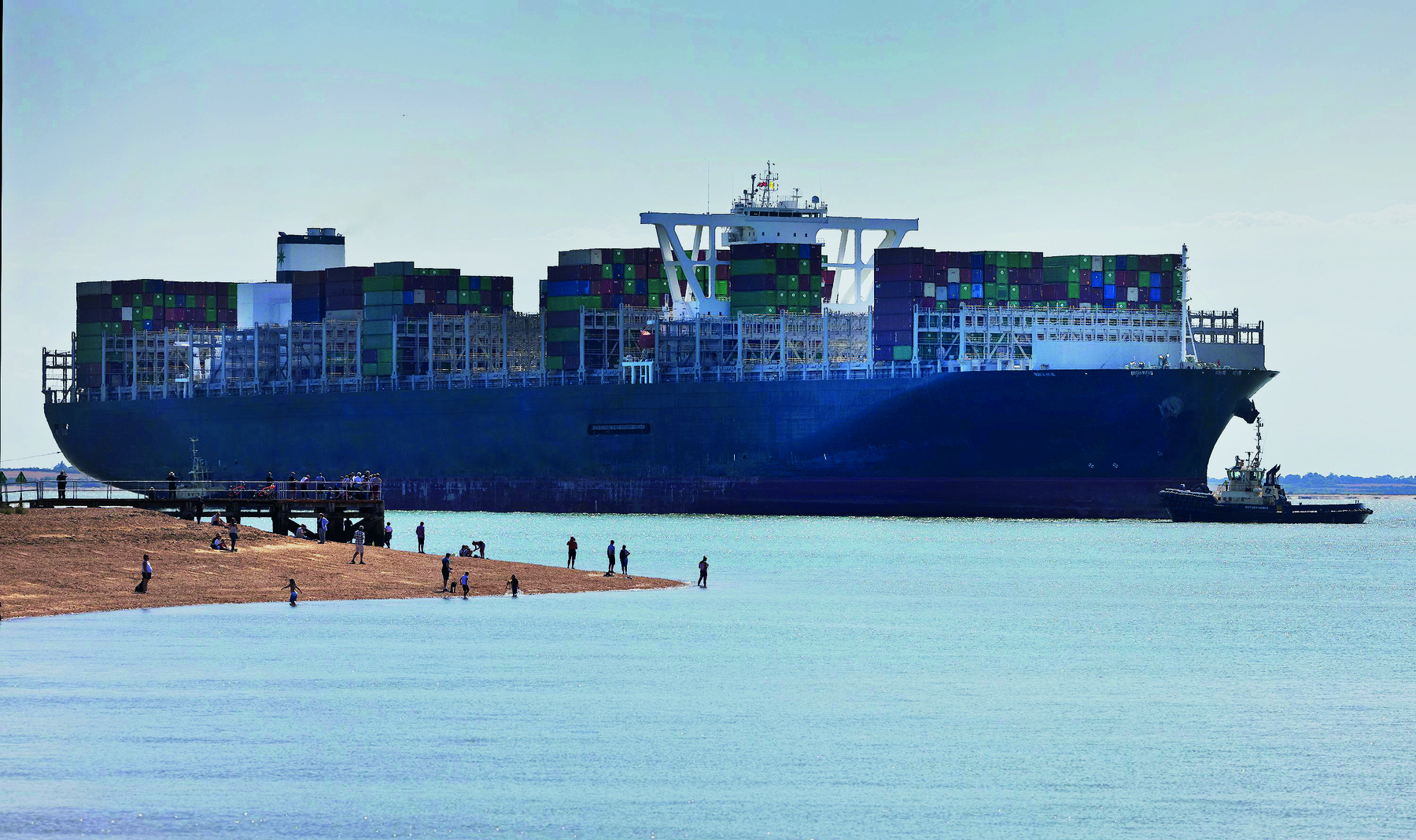 Fotografia. Vista de um grande navio cargueiro atravessando um canal marítimo. Ele carrega diversos contêineres empilhados.