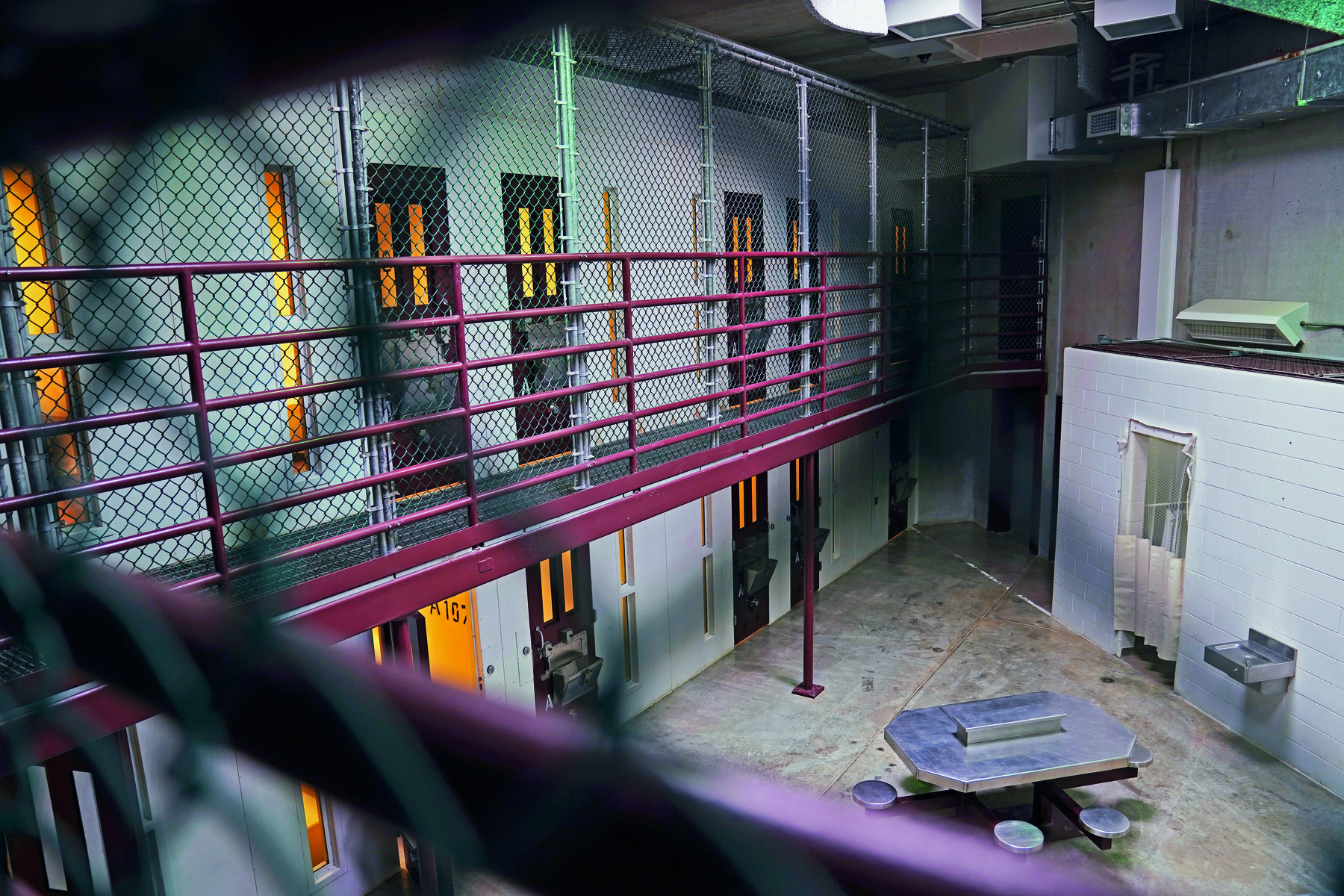 Fotografia. Pátio interno de uma prisão. São dois andares com paredes cinzentas, portas e janelas finas e compridas. O andar de cima tem grades de proteção. No pátio, uma mesa com bancos fixos.