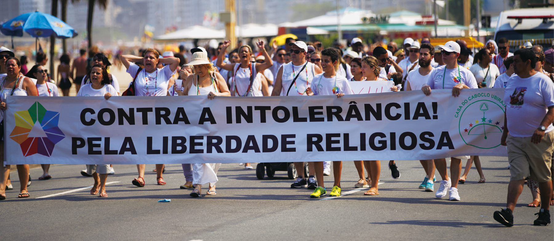 Fotografia. Pessoas em uma caminhada na rua. Elas estão vestidas de branco e carregam uma grande faixa escrito: CONTRA A INTOLERÂNCIA! PELA LIBERDADE RELIGIOSA.