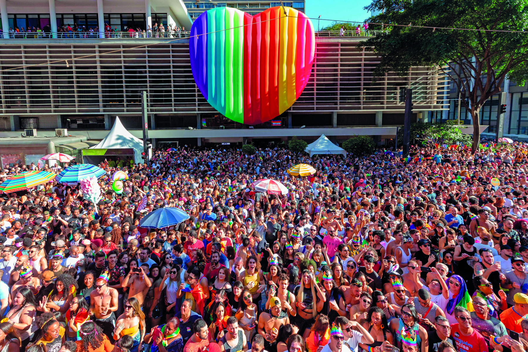 Fotografia. Multidão com adereços coloridos em desfile do Orgulho LGBTQIA+. Ao fundo, um enorme coração colorido inflável pendurado em um prédio horizontal.