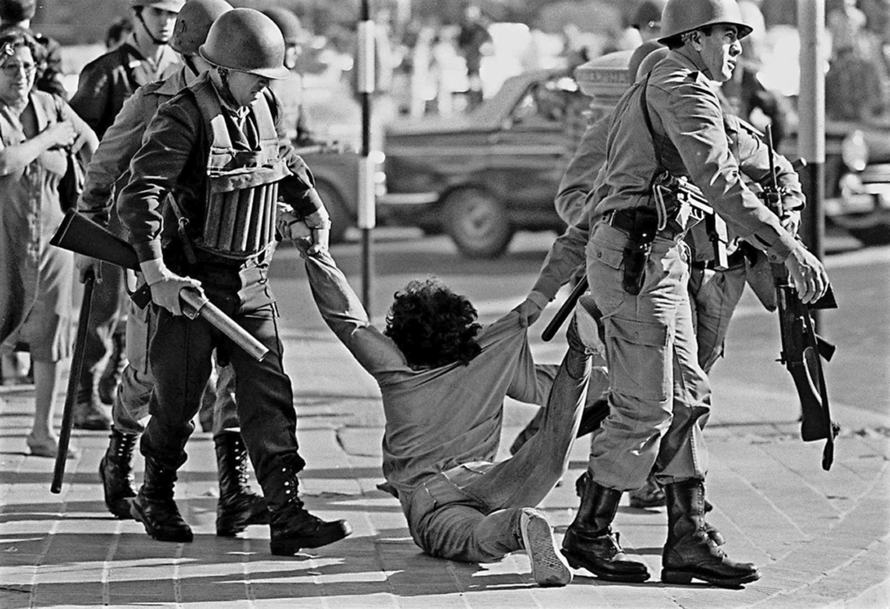 Fotografia em preto e branco. Militares arrastando uma pessoa pela rua. Eles carregam armas. Ao fundo, outras pessoas observam a cena.