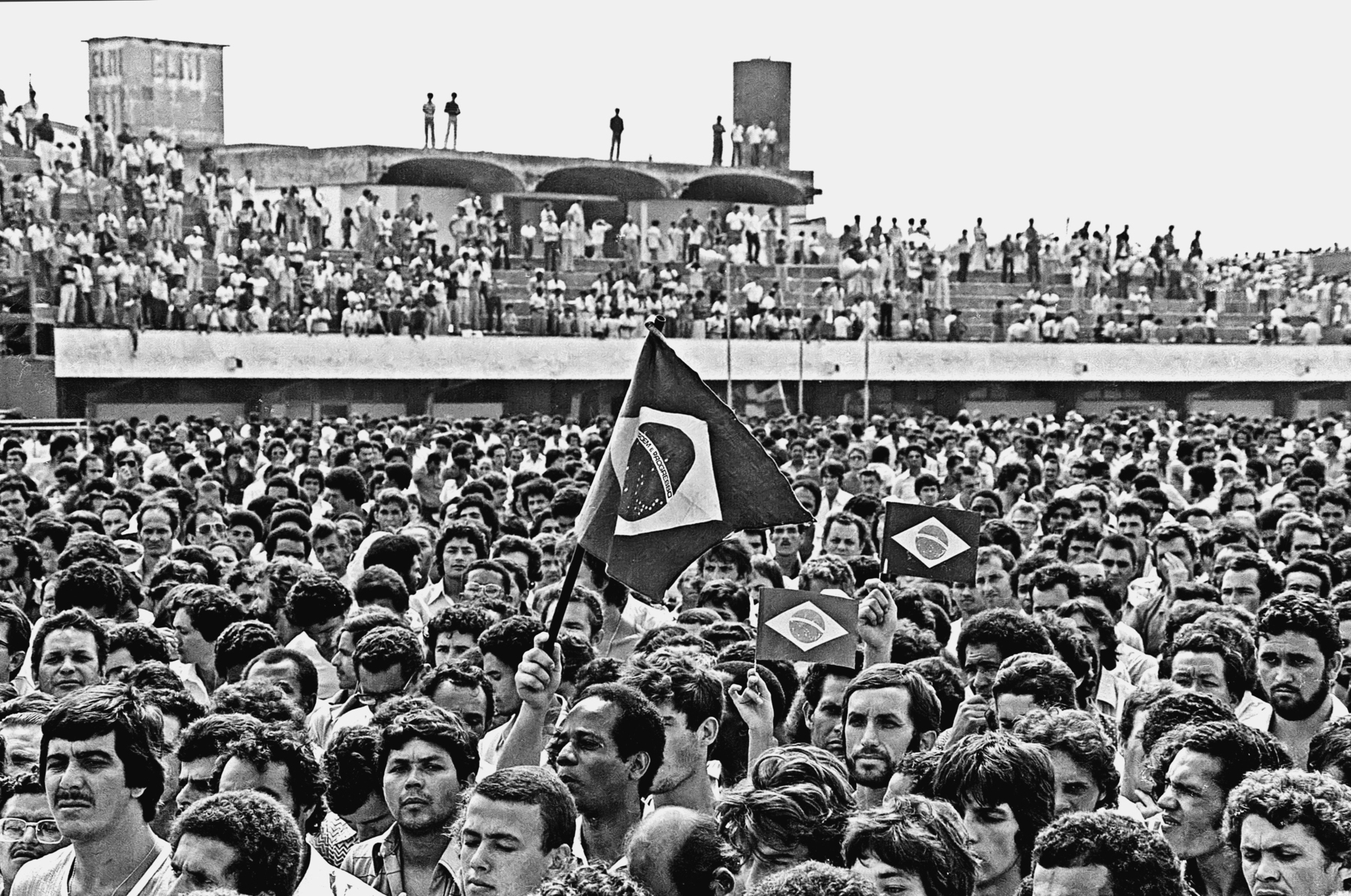 Fotografia em preto e branco. Pessoas aglomeradas em uma assembleia realizada em um estádio de futebol. Algumas carregam pequenas bandeiras do Brasil. Ao fundo, pessoas nas arquibancadas.