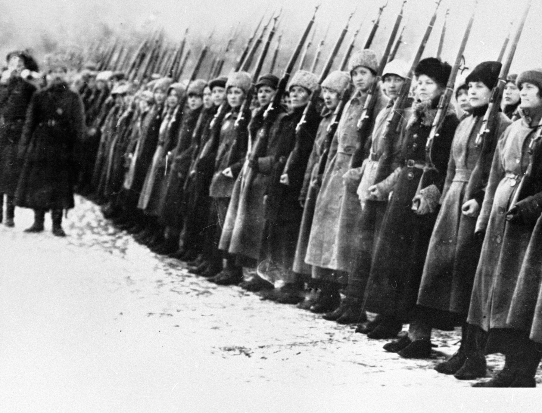 Fotografia em preto e branco. Mulheres de sobretudo e gorro, perfiladas empunhando armas de fogo com cano longo.