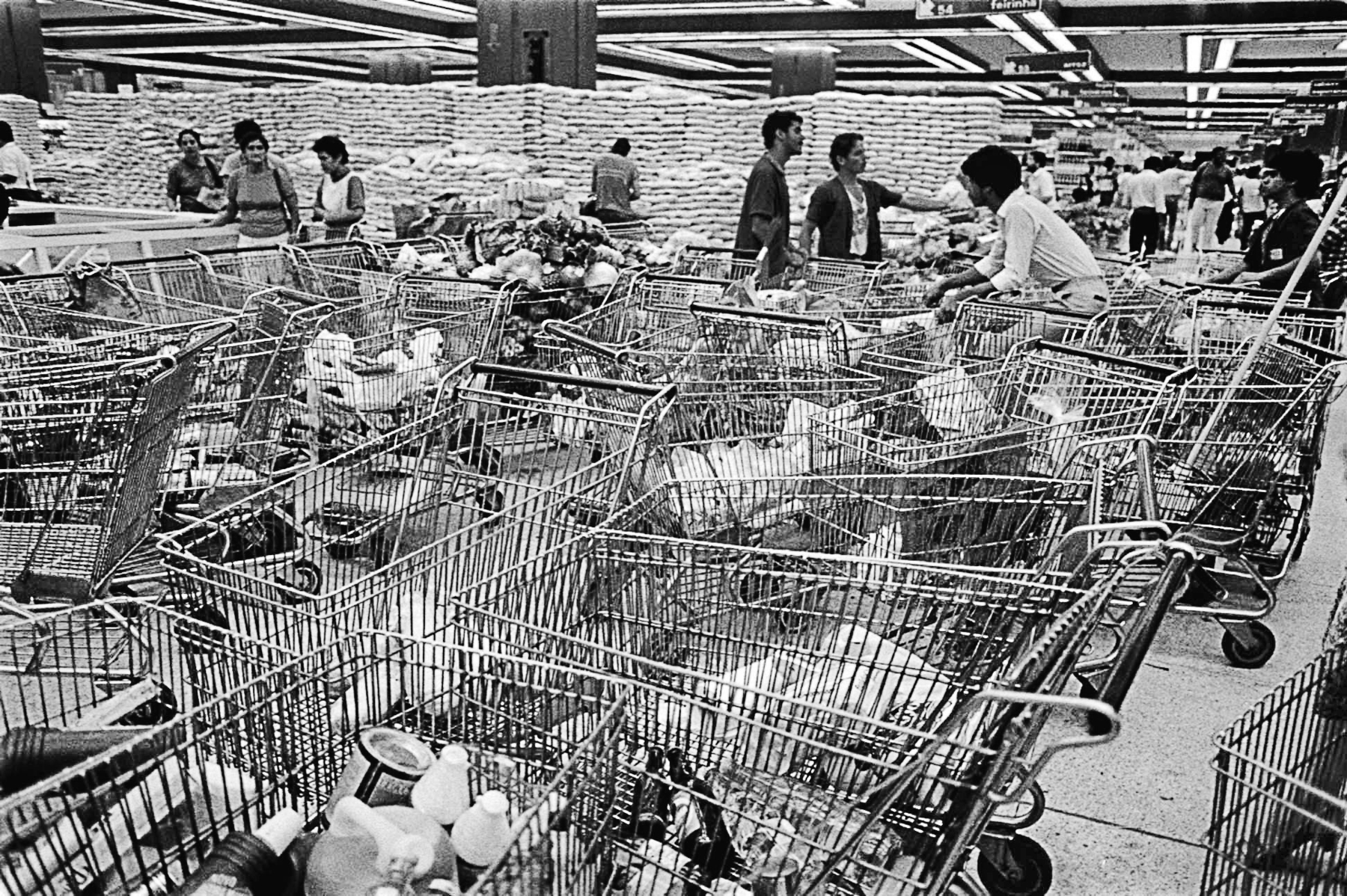 Fotografia em preto e branco. Várias pessoas dentro de um supermercado. À frente, carrinhos com compras aglomeradas.