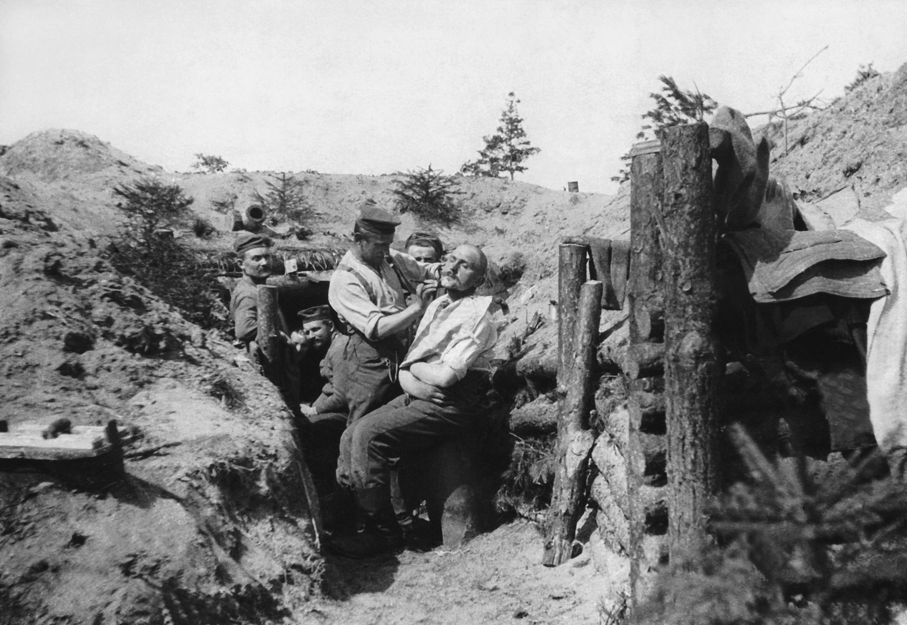 Fotografia em preto e branco. Homens dentro das trincheiras. Um deles está aparando a barba do outro, que está sentado. Ao redor, estruturas com toras de madeira na base das trincheiras.