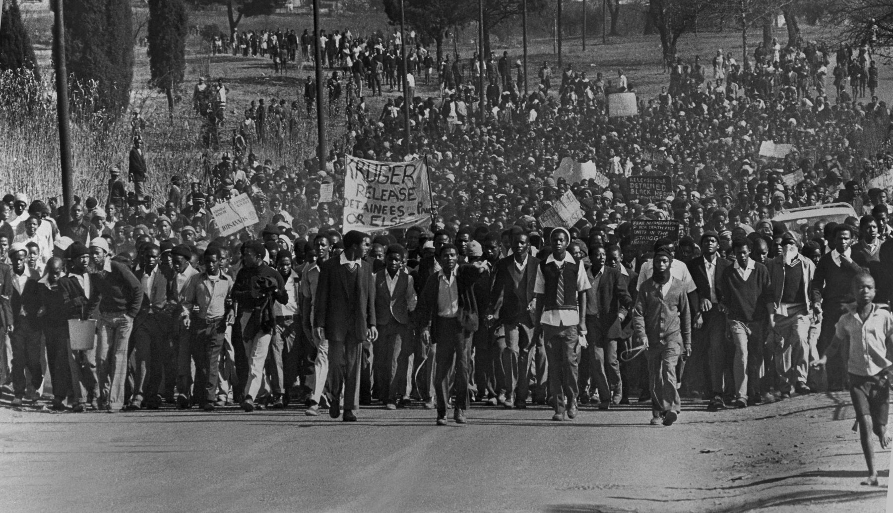 Fotografia em preto e branco. Manifestação de estudantes. Multidão em uma rua larga carregando cartazes.